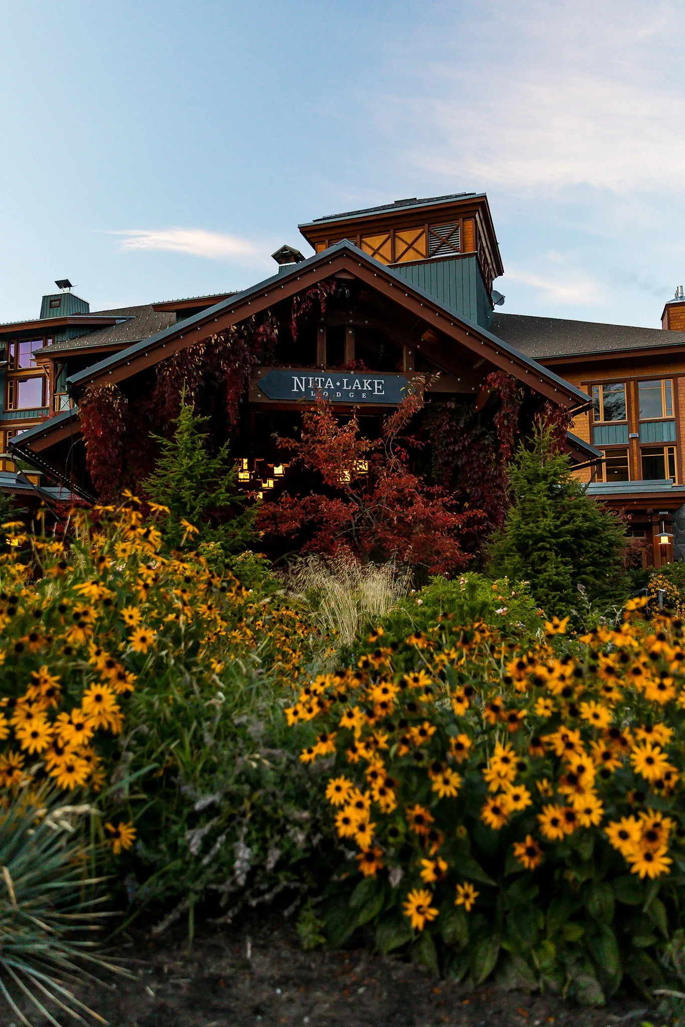 View of Nita Lake Lodge with surrounding garden of yellow flowers, trees with autumn leaves, and a partly cloudy sky.