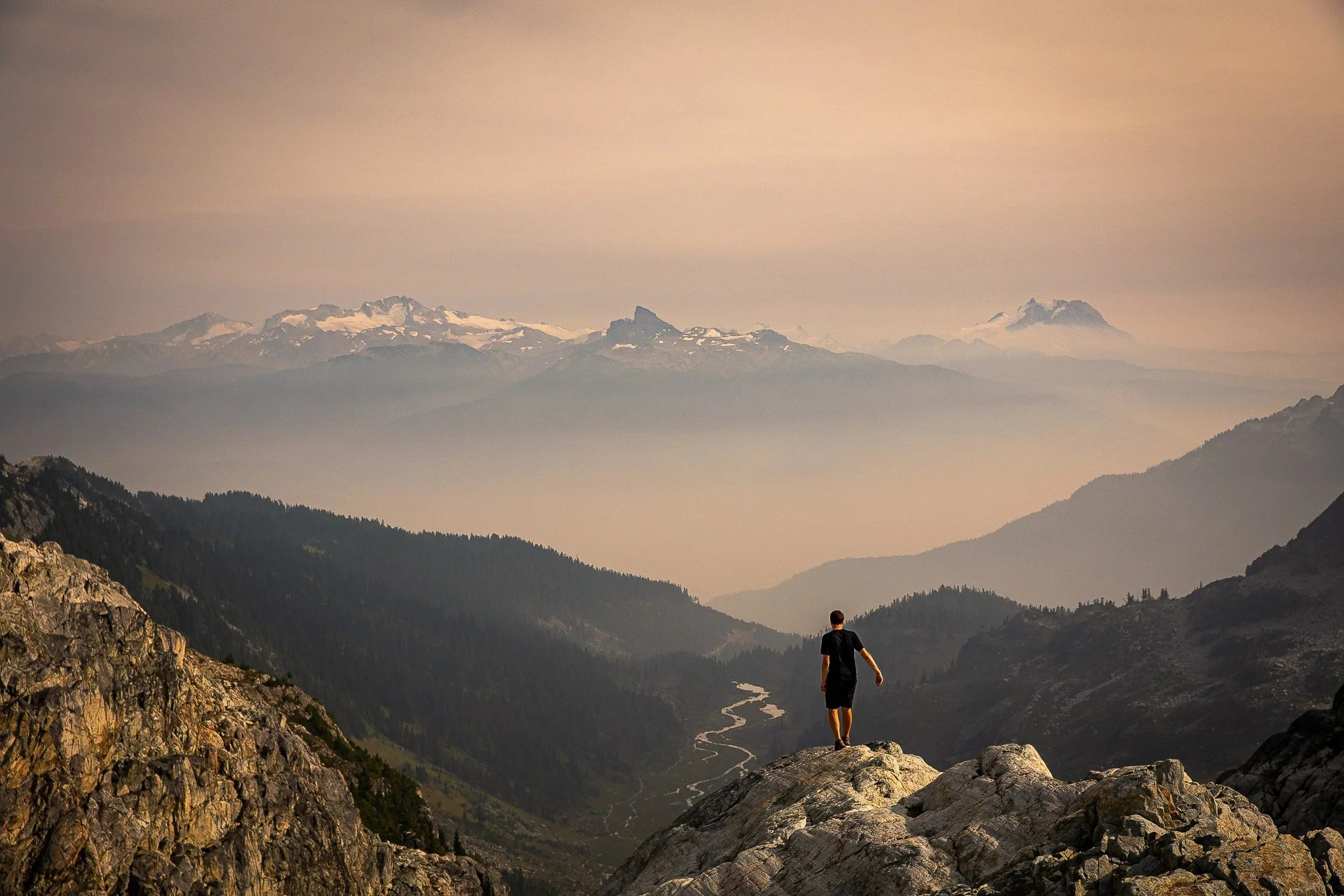 Person standing on a rocky ledge overlooking a valley with mountains in the distance and a river flowing through the valley.