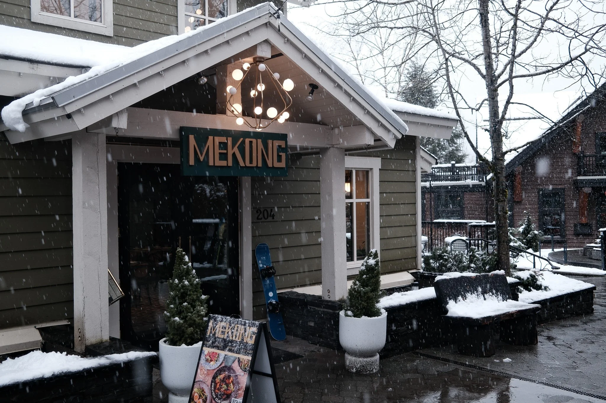 Snow-covered restaurant entrance with a sign that reads 'Mekong', potted plants, a menu board, and snow falling on the outdoor seating area.