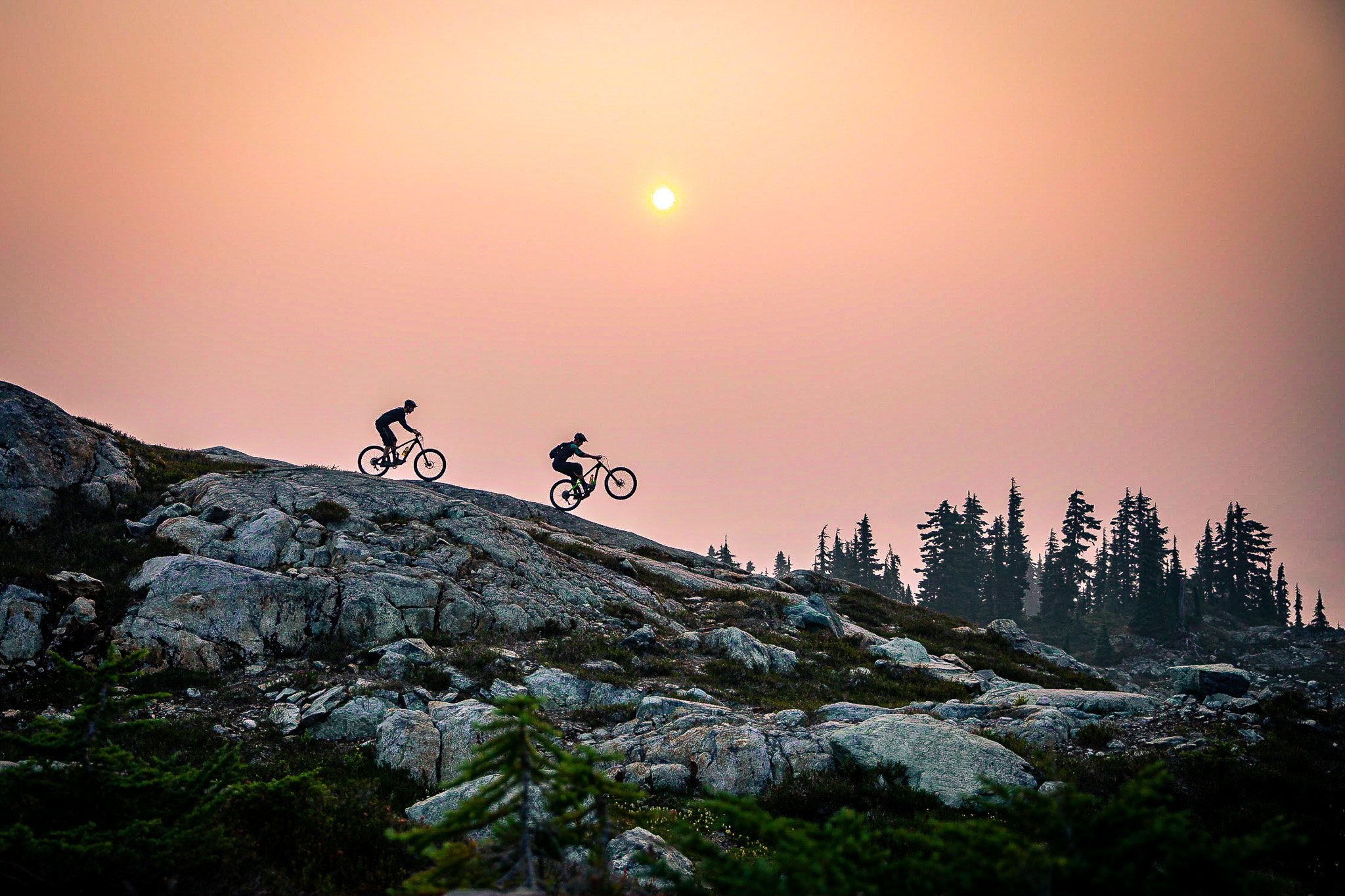Two mountain bikers riding lord of the squirells with a sunset in the background and trees in the distance.