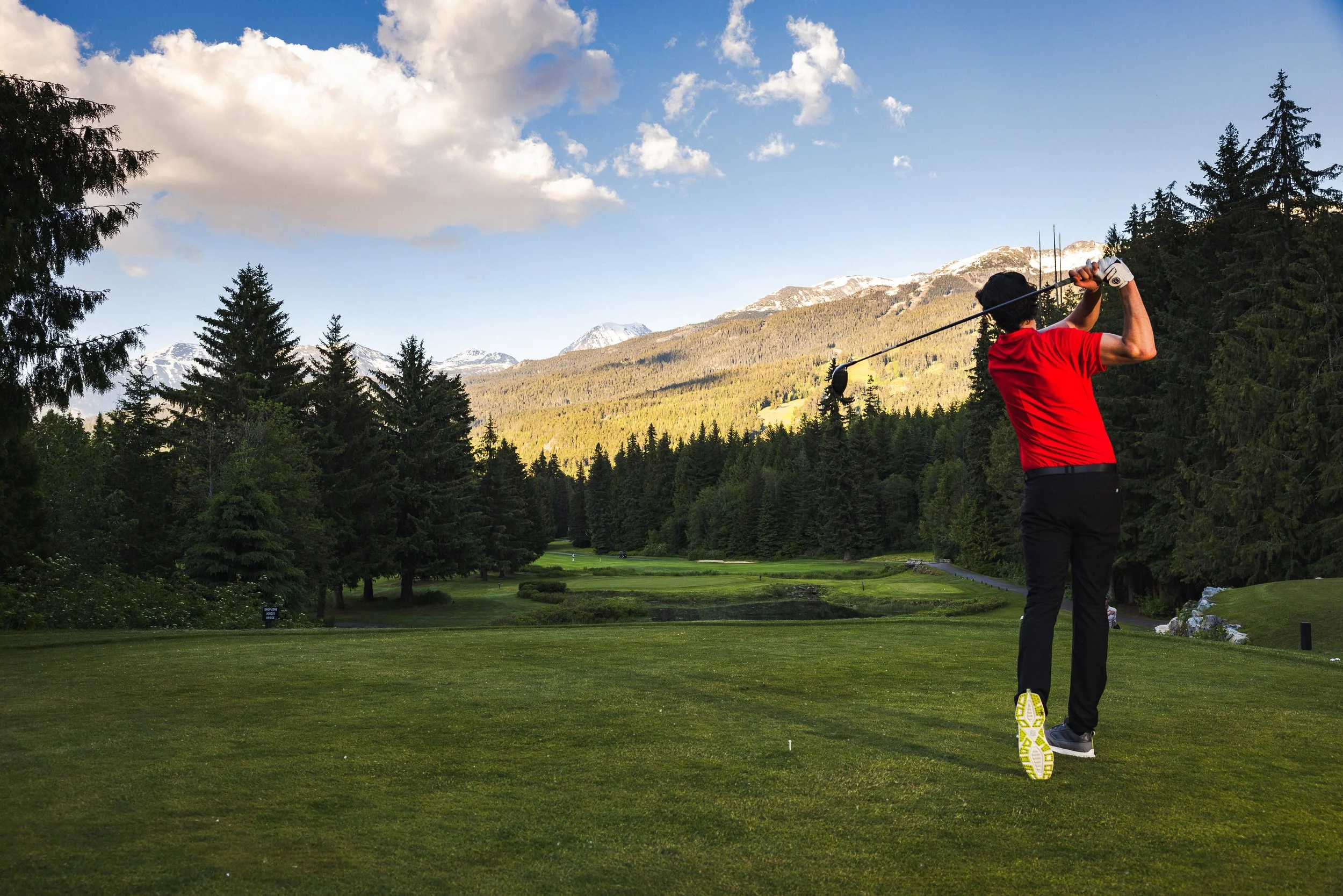 A person in a red shirt and black pants swinging a golf club on a golf course surrounded by trees and mountains.