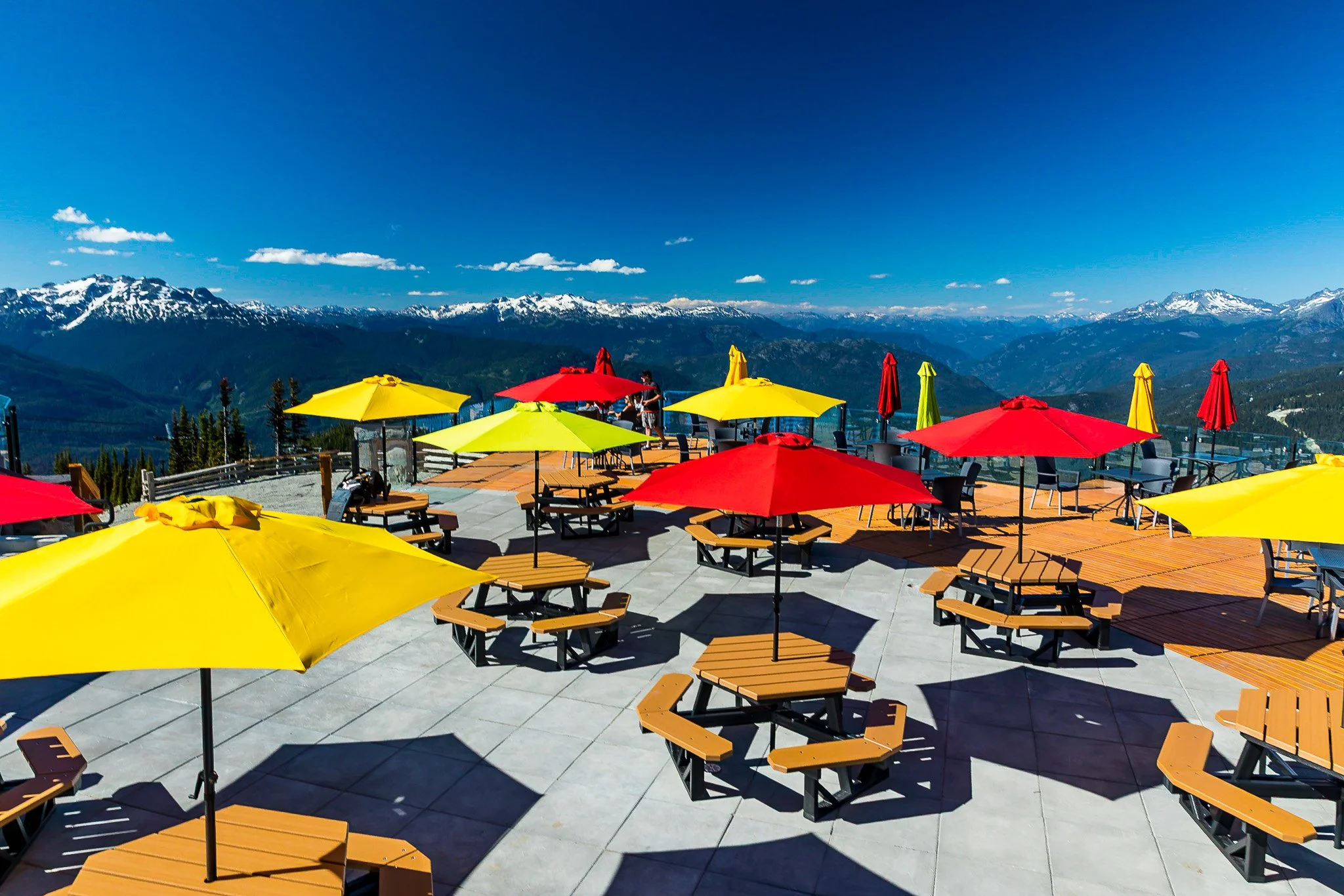 Outdoor seating area with yellow and red umbrellas, wooden picnic tables, and mountain view with snow-capped peaks under a blue sky.