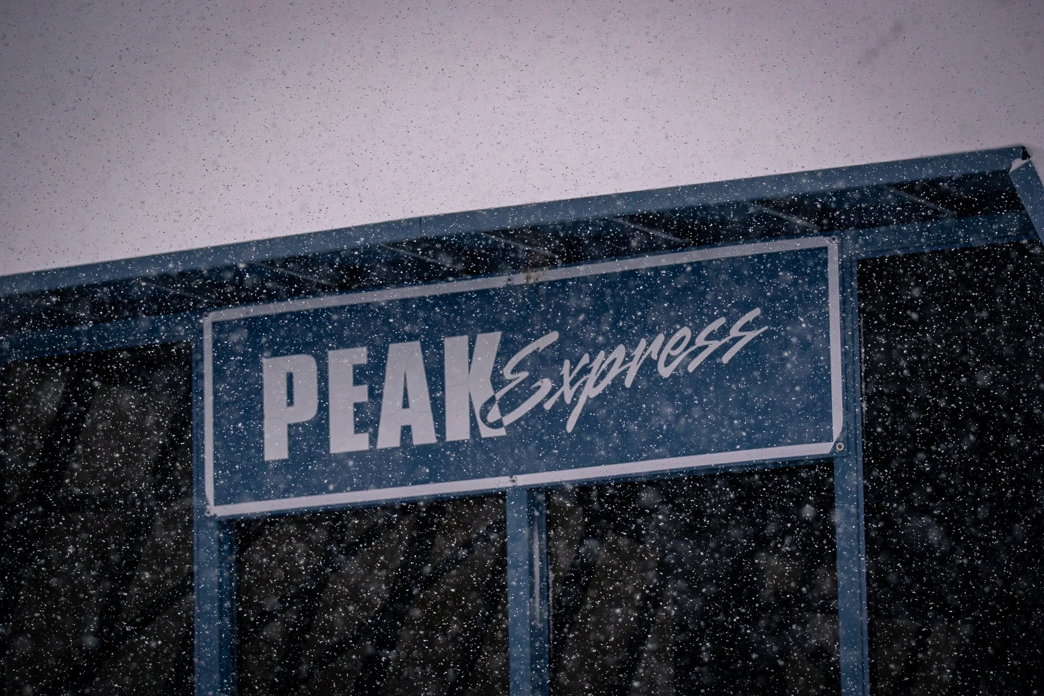 A blue sign with white text that reads 'PEAK express' is seen through snowfall against a grey sky.
