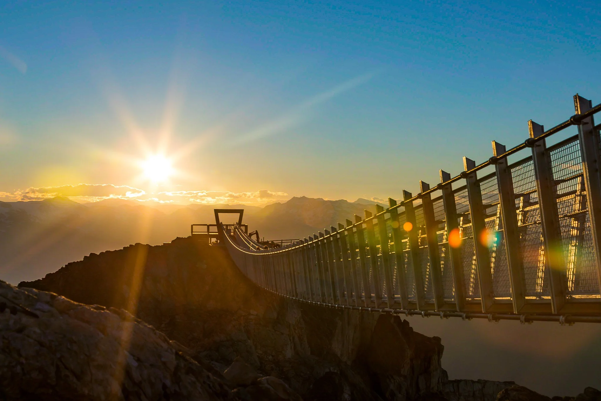 Suspension bridge on a mountain at sunset with the sun shining brightly in the sky.