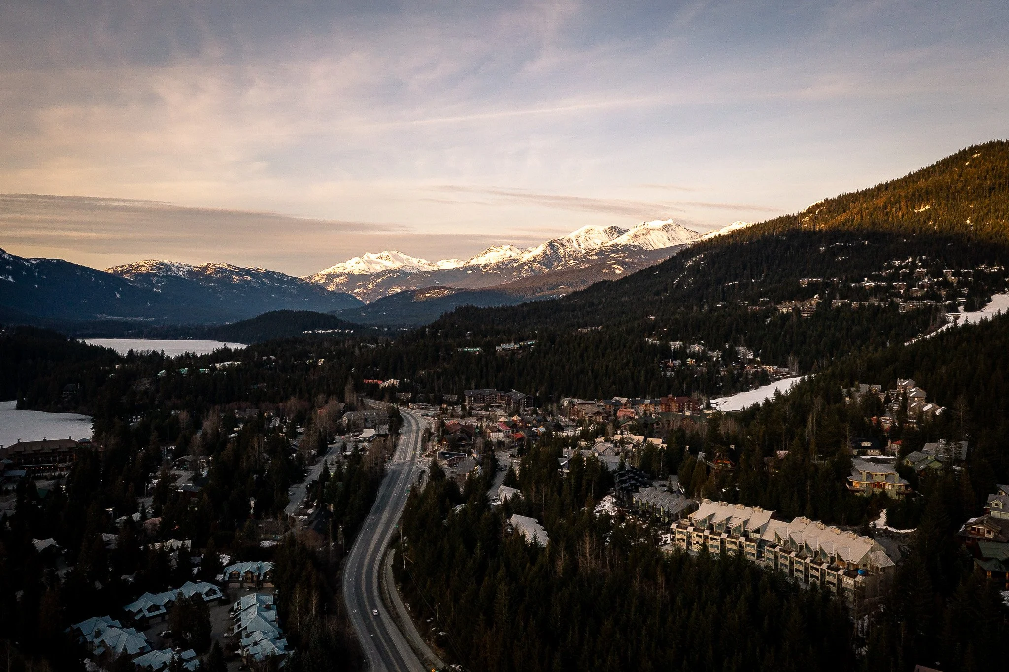 Aerial view of a mountain town surrounded by snow-capped mountains and dense forest, with a winding road running through the town and buildings on either side.