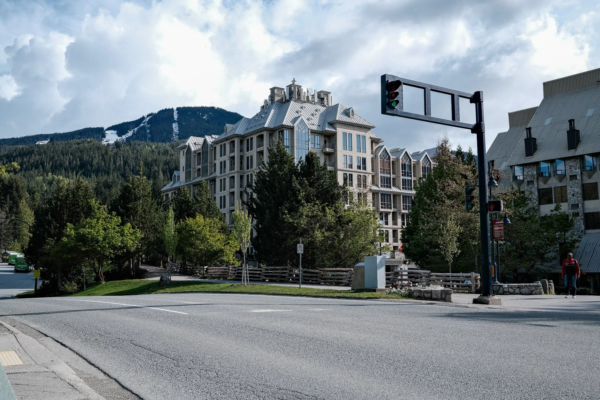A street scene with a large, modern building in the background, trees, a traffic light, and a person walking along the sidewalk.