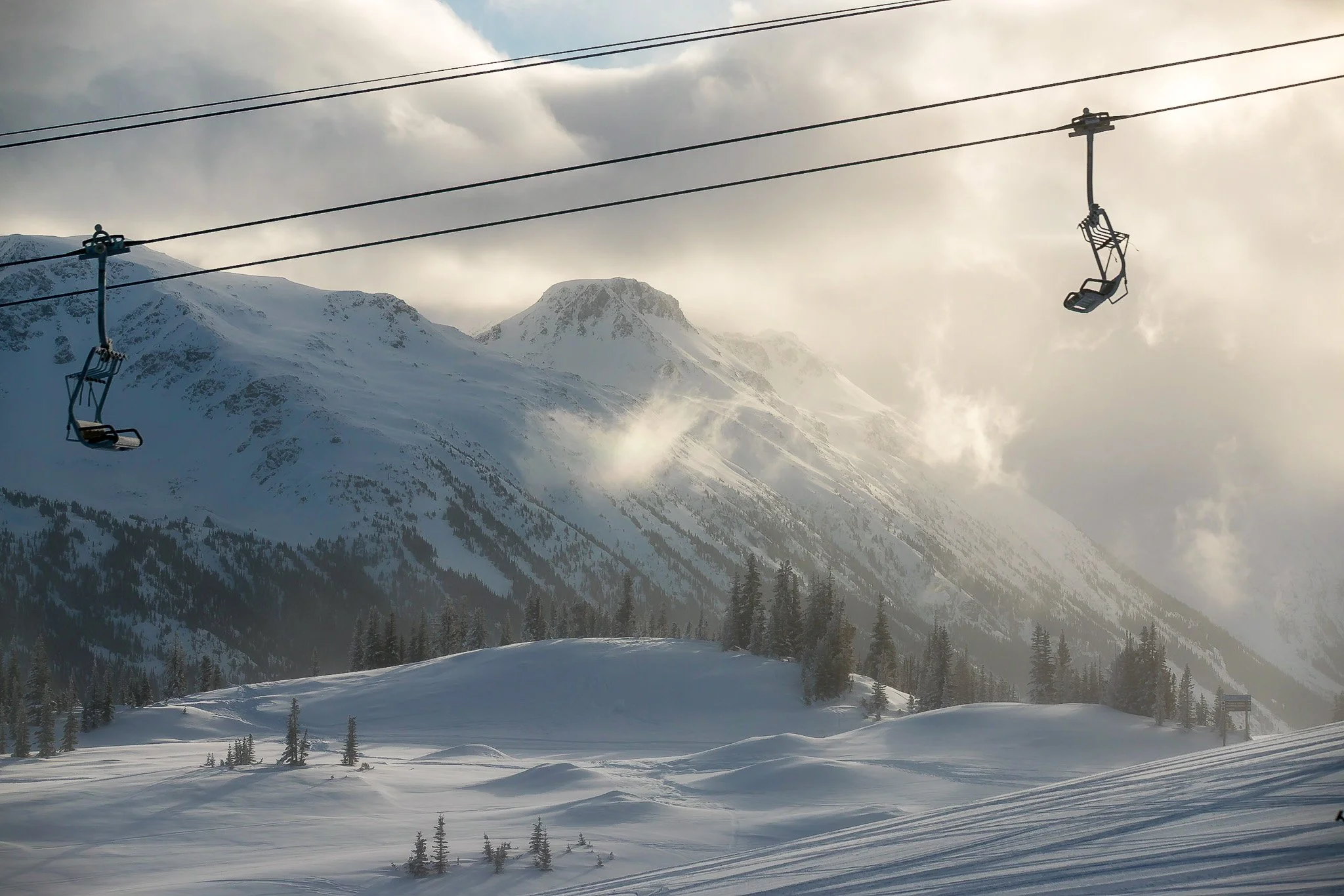Snow-covered mountain landscape with ski lift chairs hanging from cable, and pine trees in foreground.