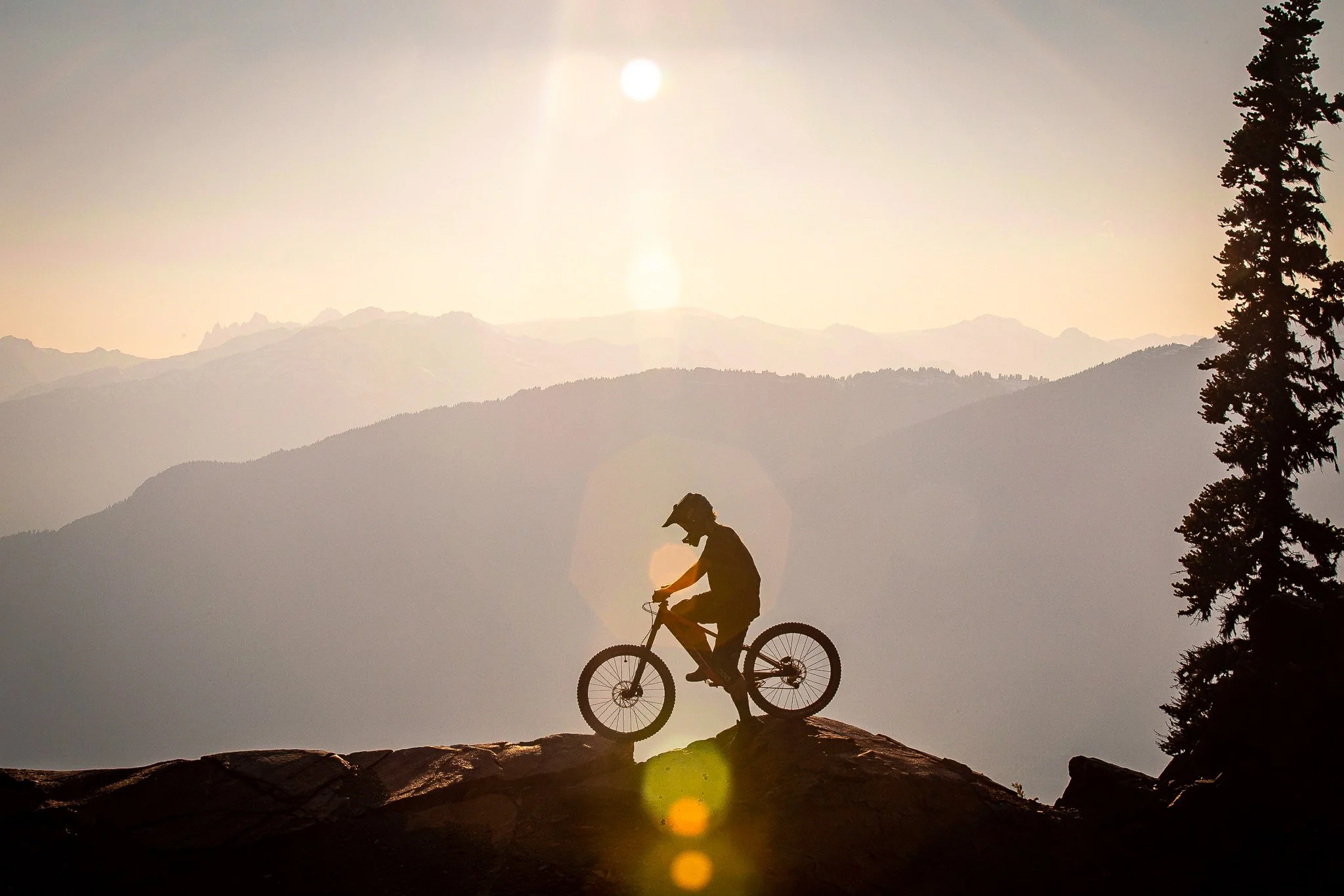 Silhouette of a person riding a mountain bike on a rocky trail during sunset, with mountains in the background and a sun casting a glow.