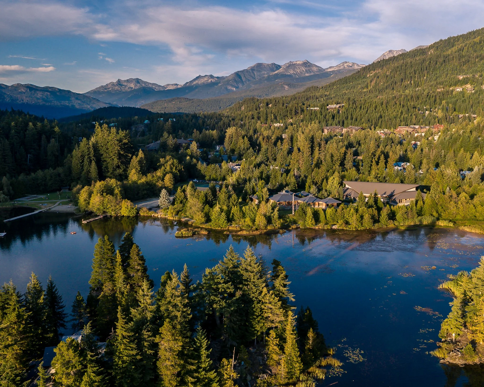 Aerial view of a forested landscape with a lake, houses, and mountains in the background under a partly cloudy sky.