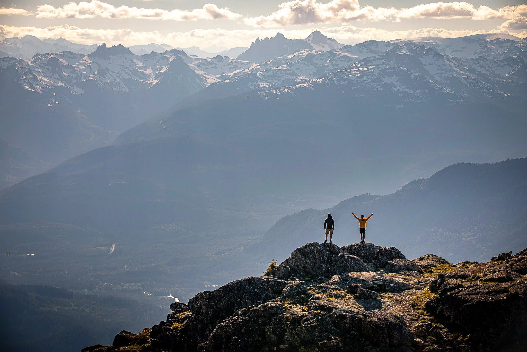 Two hikers standing on a rocky ledge overlooking a mountain range with snow-capped peaks, one with arms raised in celebration.