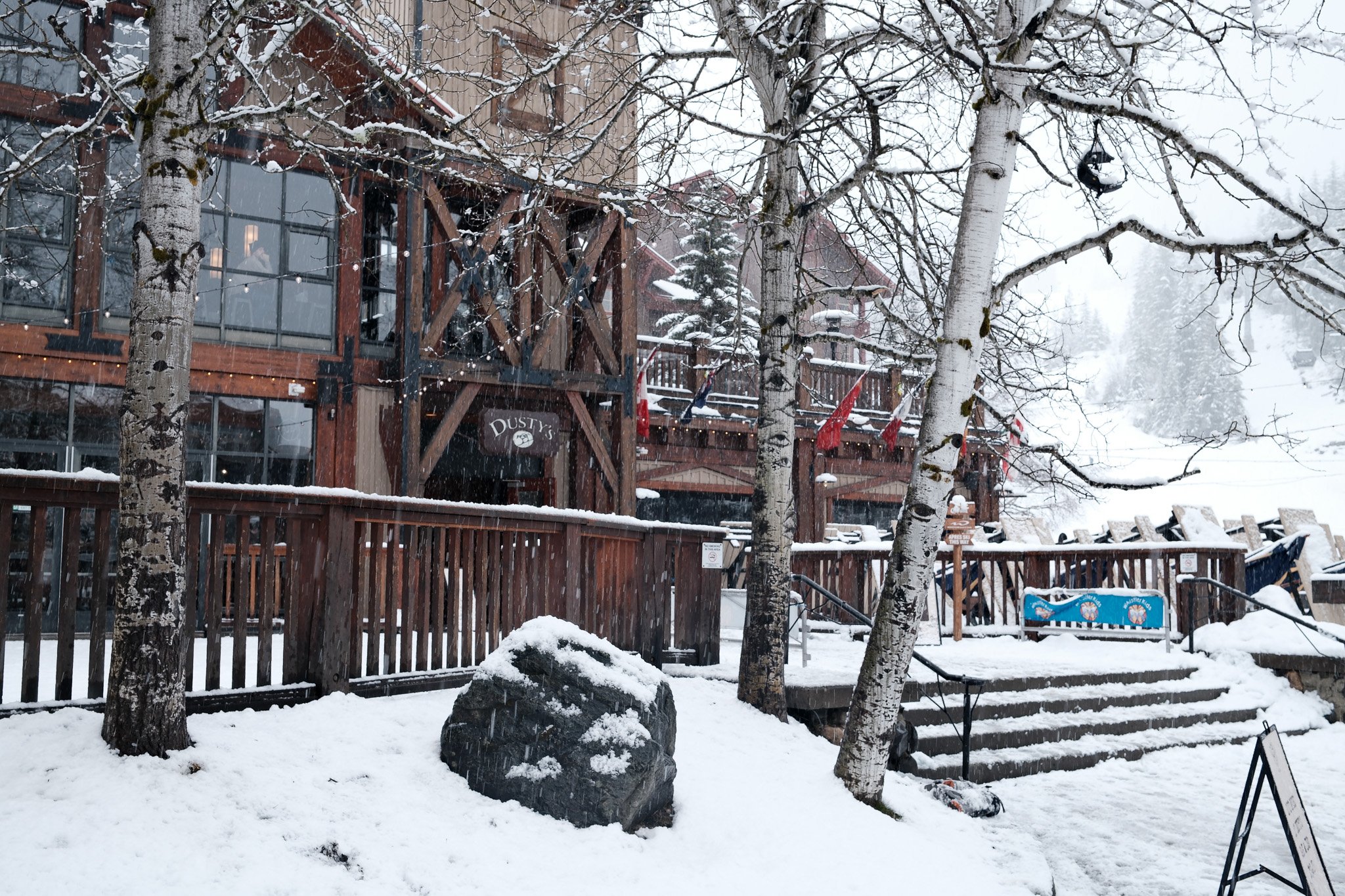 A snowy scene of a rustic wooden building named 'DUSTY'S', with trees covered in snow, a rock with snow on it, and a snowy outdoor staircase leading to a deck with flags.