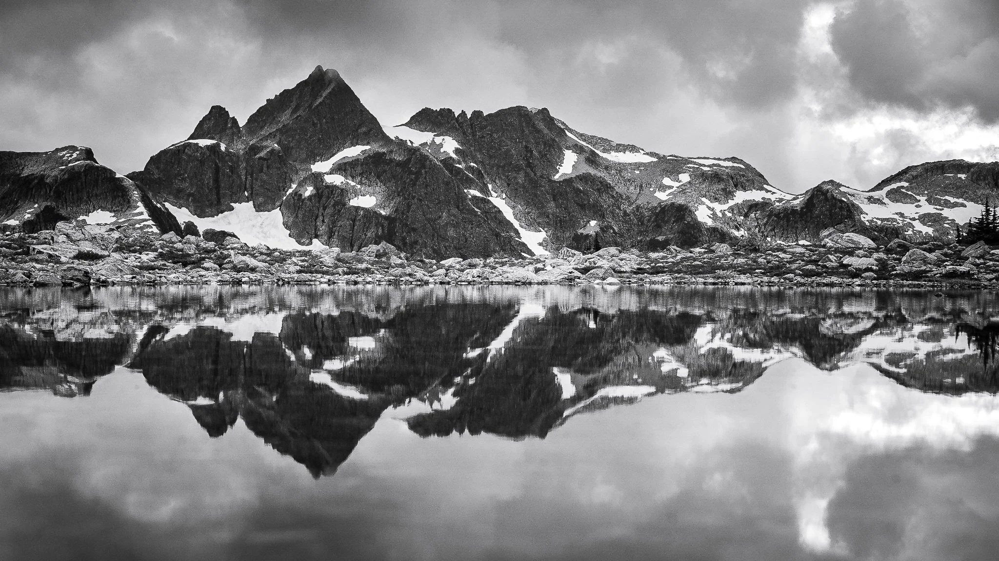 Black and white photograph of a mountain range with snow patches, reflected in a calm body of water in the foreground, under cloudy sky.