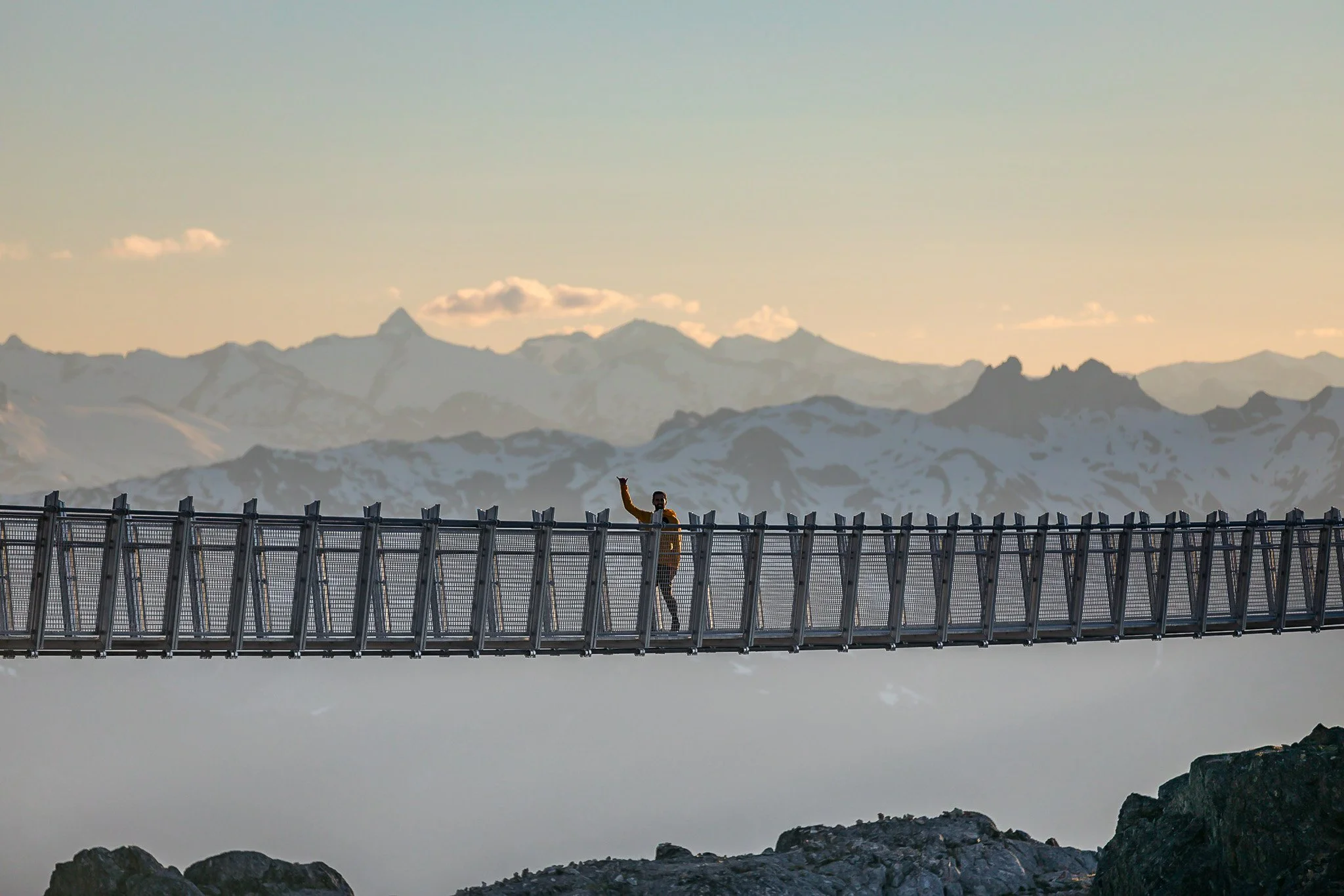 A person walking on a suspended bridge high in the mountains with snow-capped peaks in the background, waving.