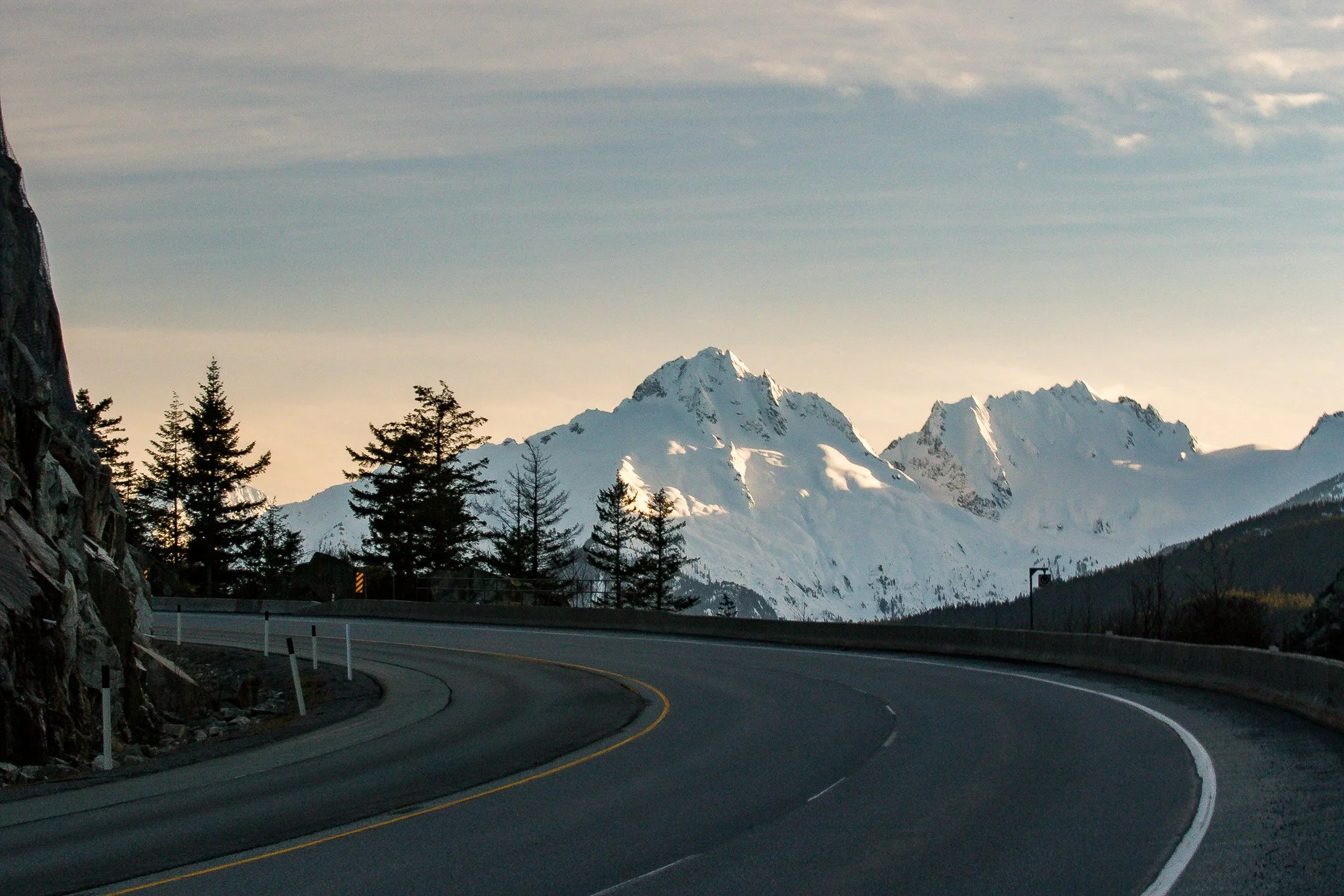 A winding mountain road with a guardrail, surrounded by trees, leading towards snow-covered peaks in the distance, under a partly cloudy sky.