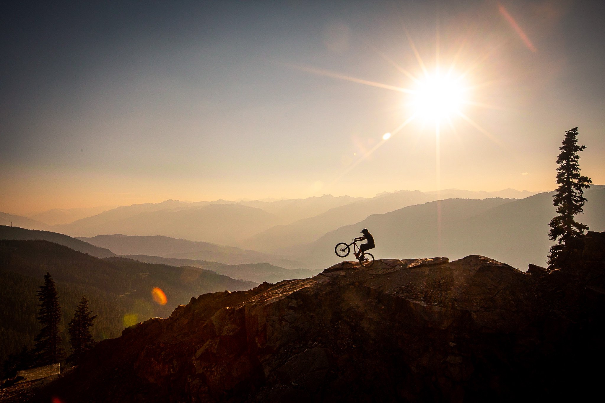 A person performing a wheelie on a mountain trail during sunset with a mountain range in the background.