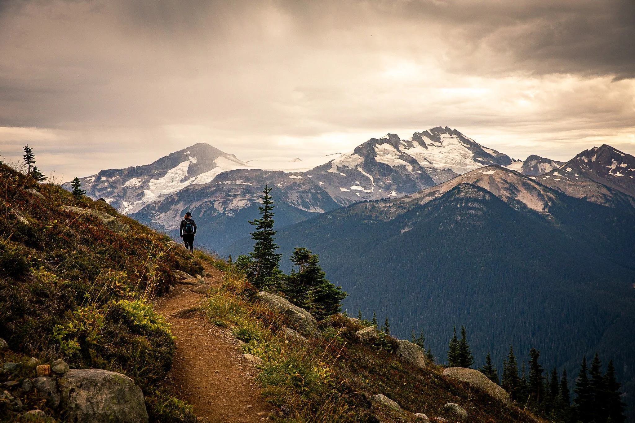 A hiker walking on a trail on a mountainside with snow-capped peaks in the background, under a cloudy sky.