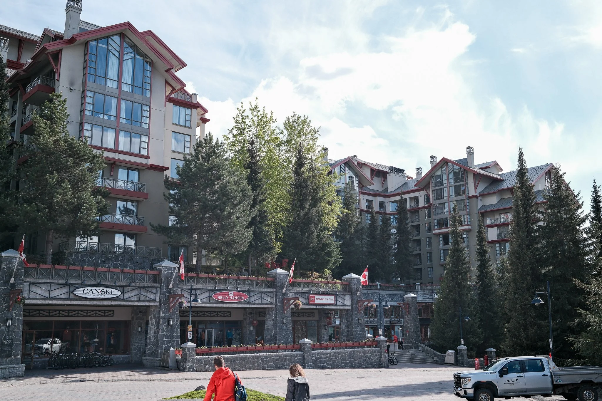 Multiple multi-story buildings with large windows and red accents, surrounded by tall trees, under a partly cloudy sky, with two people walking and a white pickup truck parked in front.