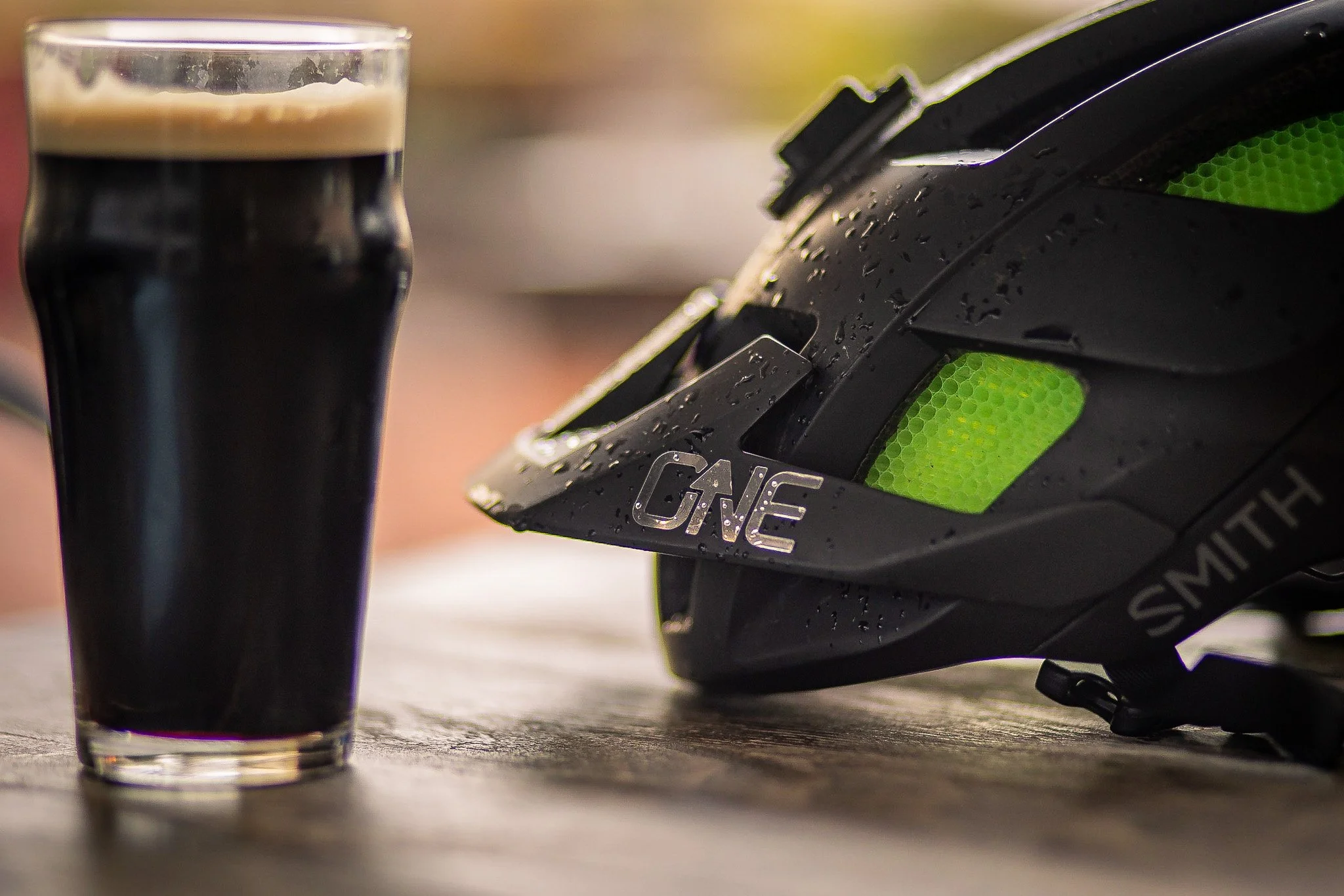 A glass of dark stout beer and a black cycling helmet with green accents resting on a wooden surface, with water droplets visible on the helmet.