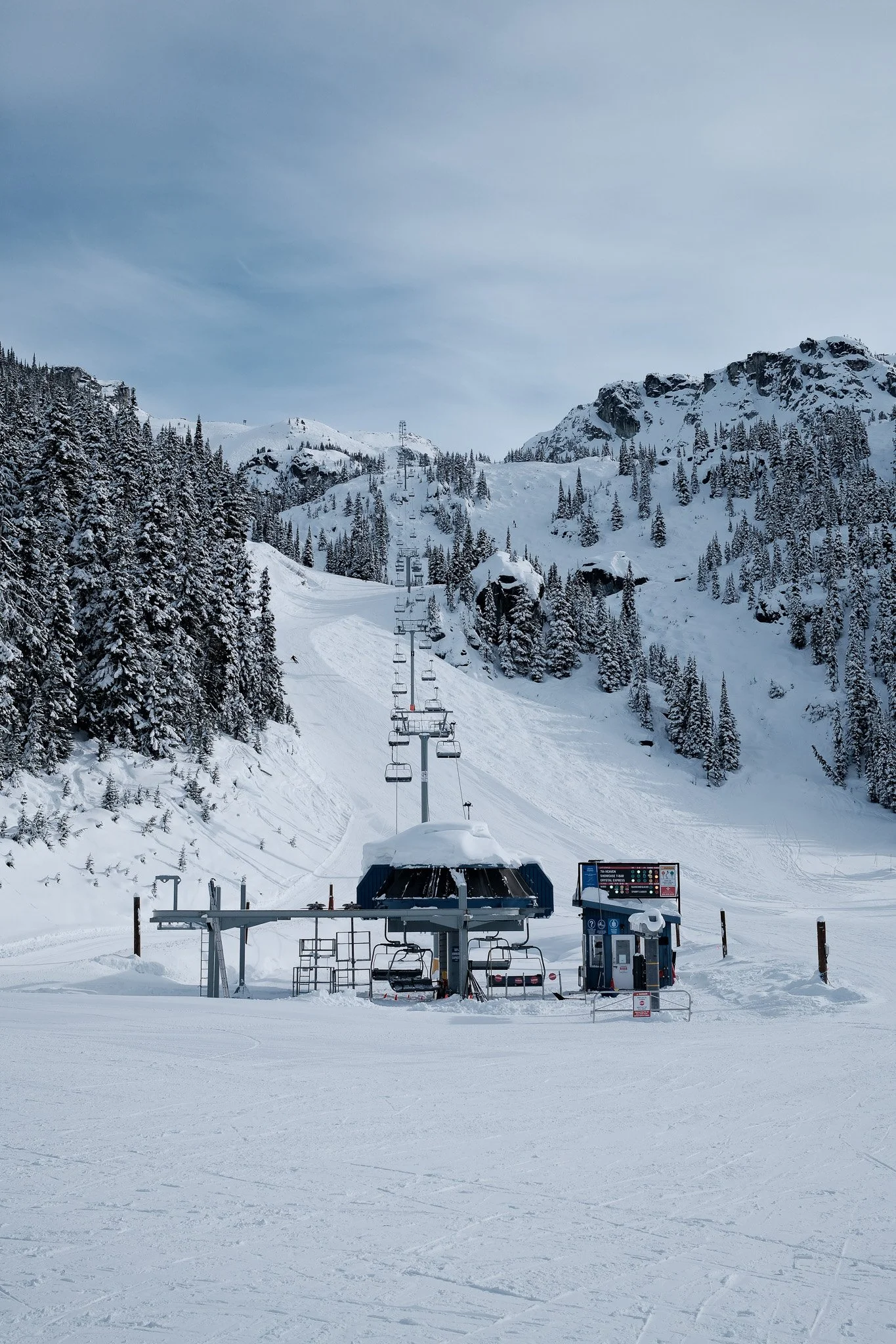 Empty ski lift at a snowy mountain ski resort with pine trees and mountain peaks in the background.