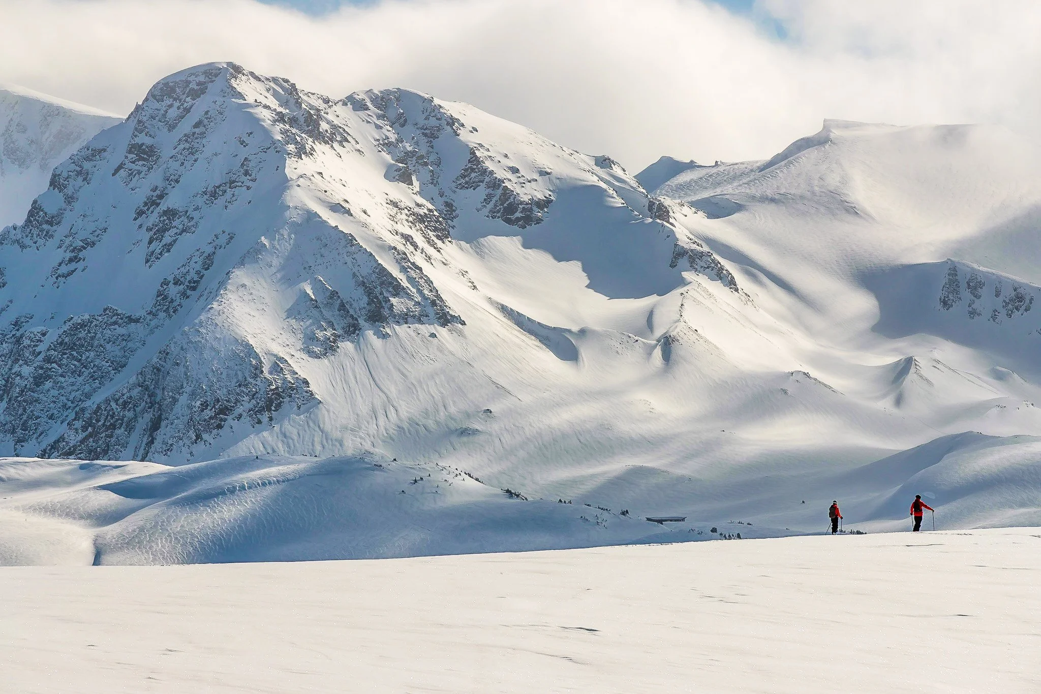 Snow-covered mountain range with two skiers in red jackets and black pants in the foreground.