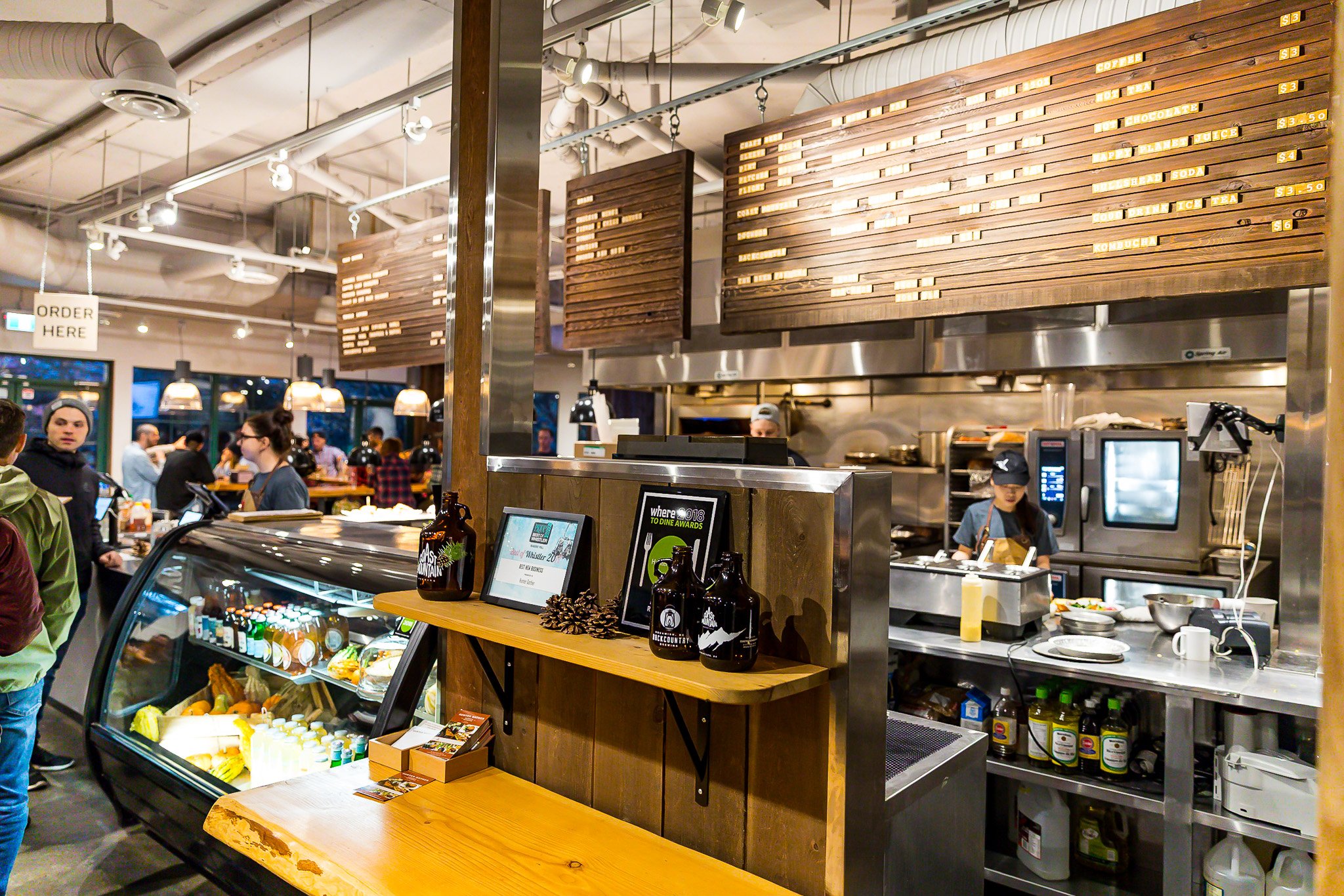 Inside a busy restaurant with a visible open kitchen area, people waiting in line, wooden menu boards, and a refrigerated display case filled with food and drinks.