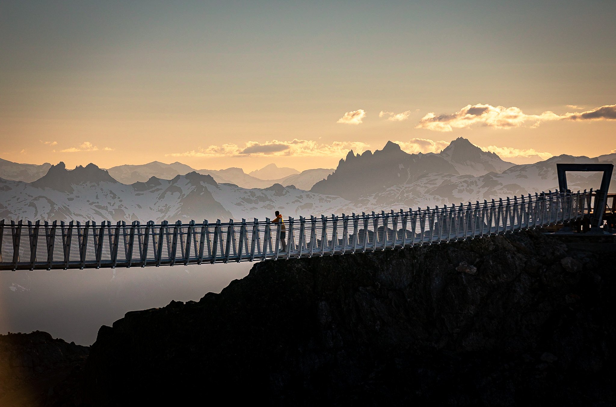 A person walking across a suspension bridge over a rocky cliff with snow-covered mountains in the background during sunset.