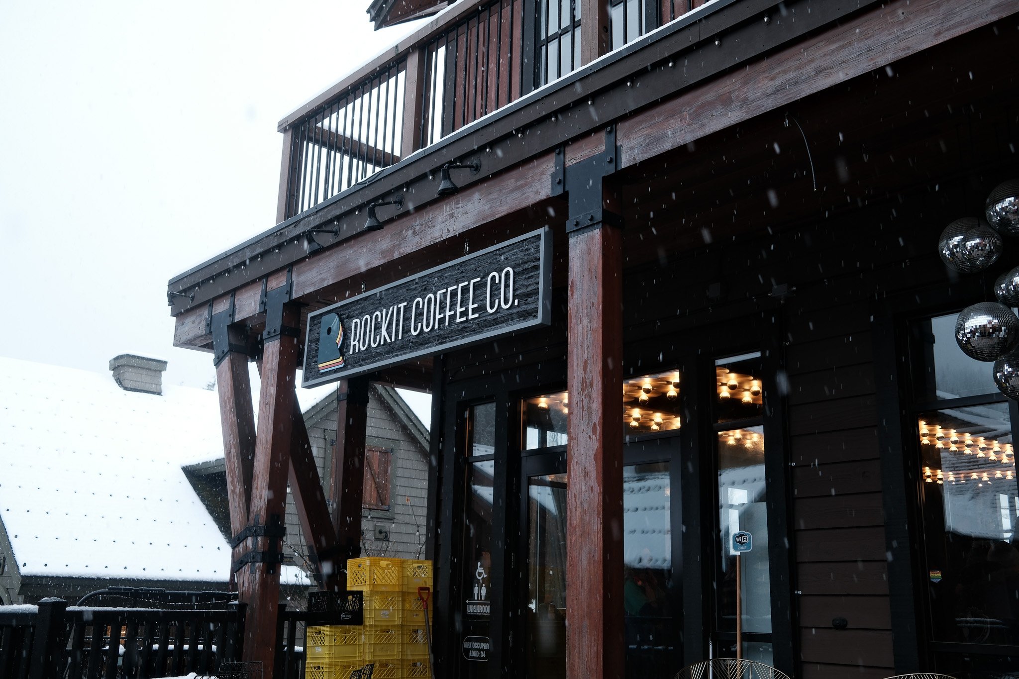 Exterior view of Rockit Coffee Co. in a snowy environment, featuring a wooden building with a balcony and a signboard, with yellow crates and snow falling.
