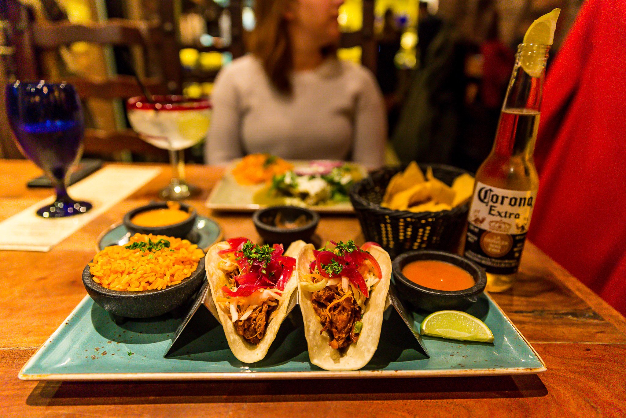 A plate of Mexican food featuring tacos filled with shredded beef, topped with red peppers and onions, alongside bowls of rice, sauces, chips, and a lime wedge on a wooden table at a restaurant.