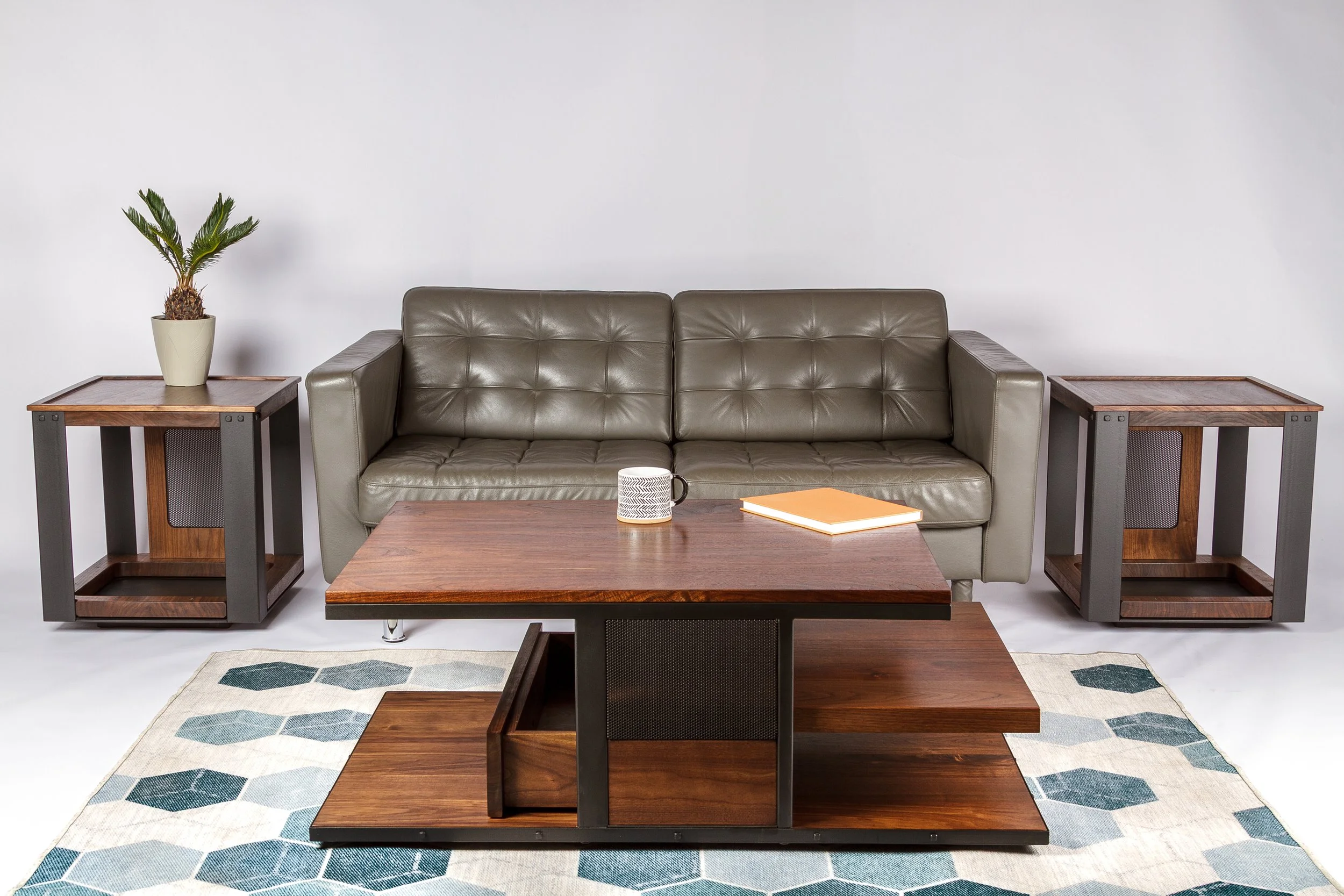 Modern living room setup with a gray leather couch, wooden coffee table, two matching side tables, a potted plant, a mug, a book, and a hexagonal-patterned rug.