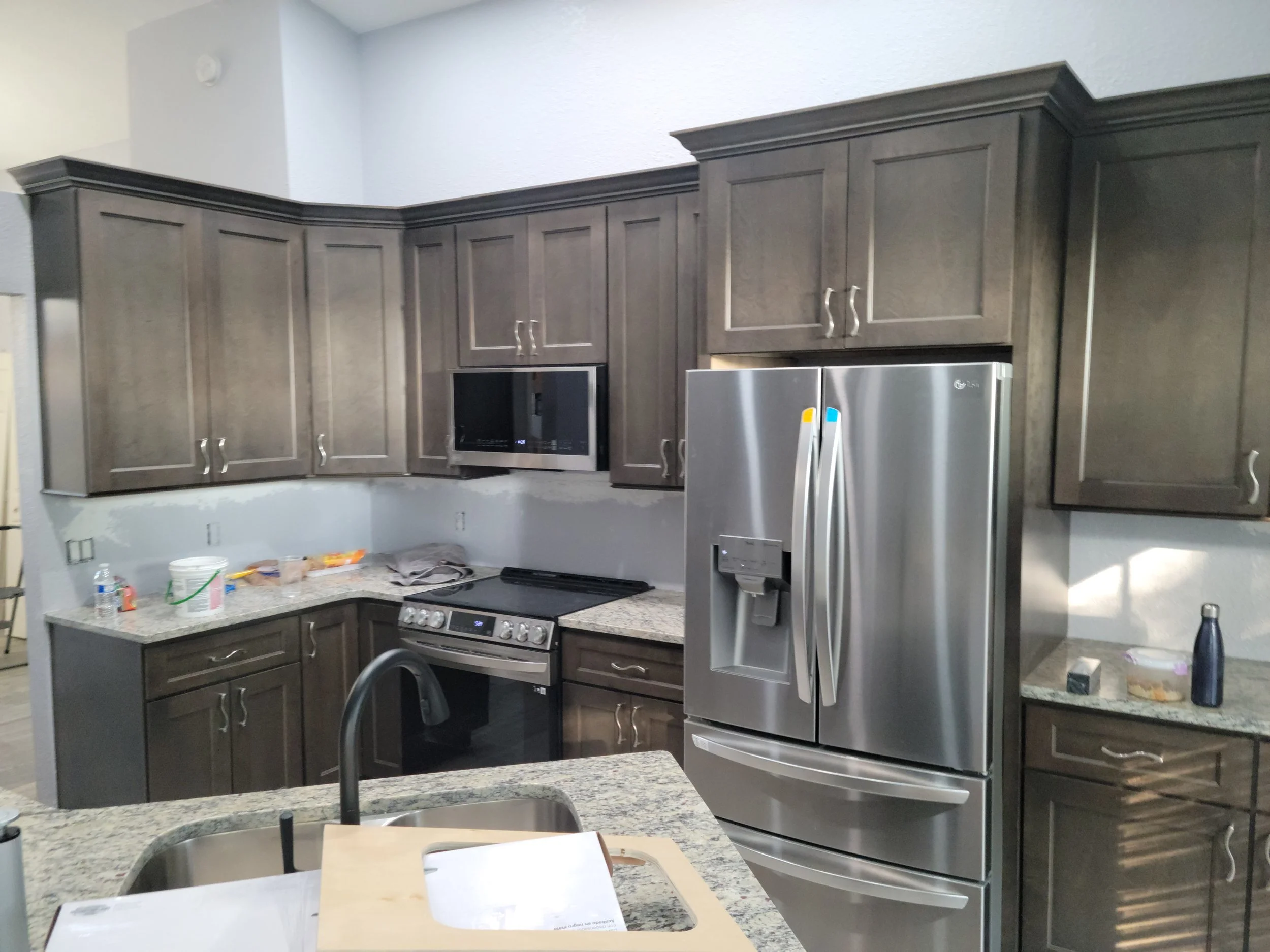 A kitchen with dark wooden cabinets, a granite countertop, stainless steel refrigerator, microwave, and stove. Various items and cleaning supplies are on the countertop.