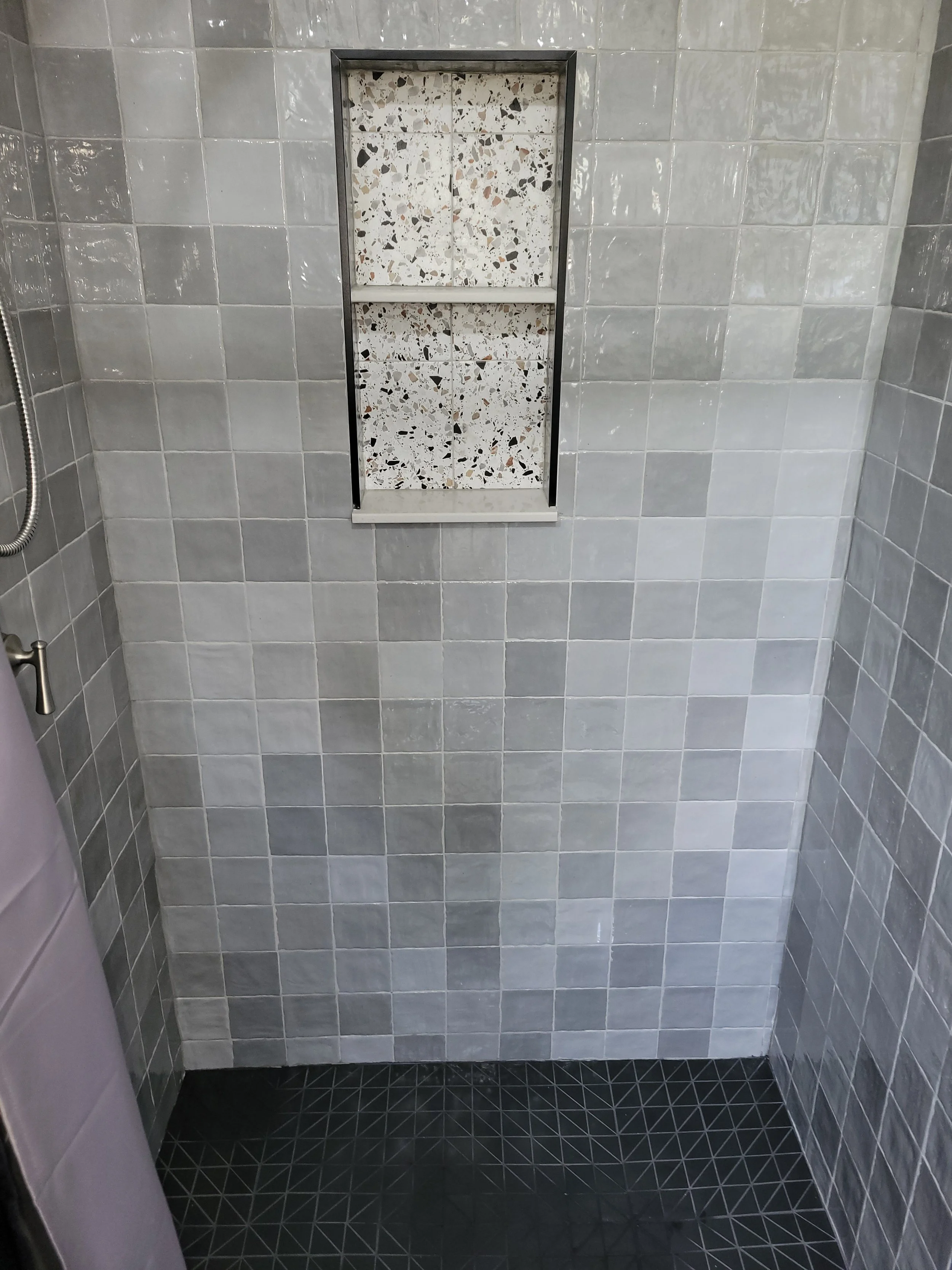 Shower with light gray square tiles, a built-in niche with terrazzo tiles, and black geometric patterned floor tiles.