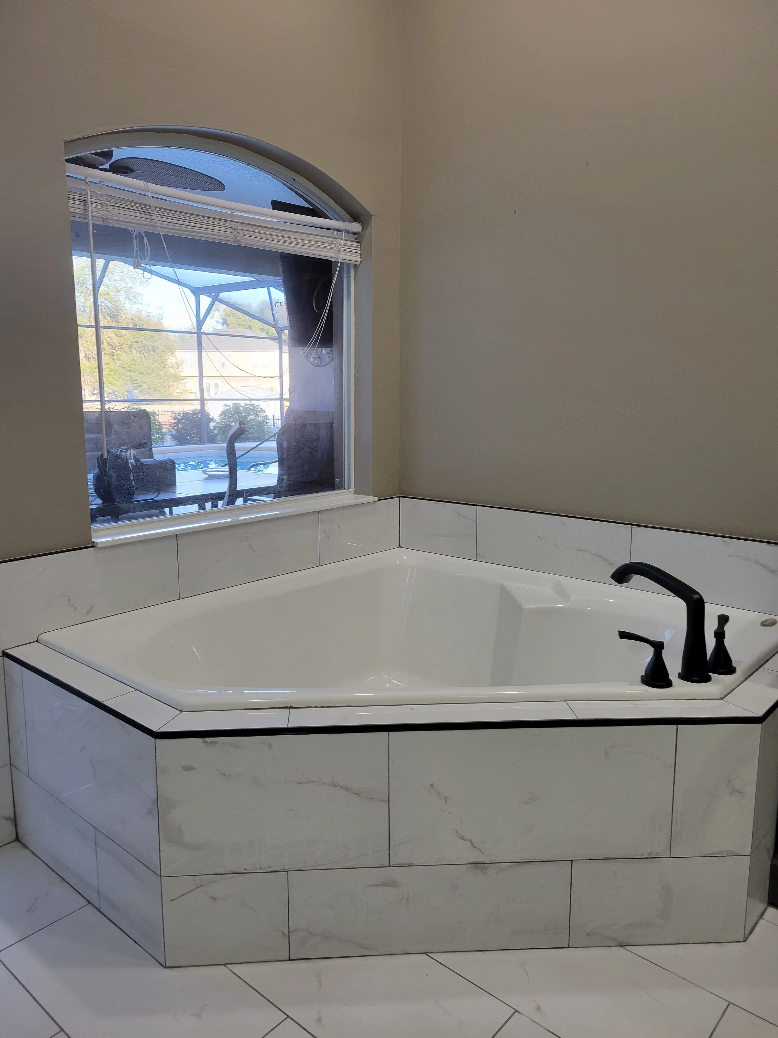 White corner bathtub with black fixtures in a bathroom next to a window showing an outdoor patio and trees.