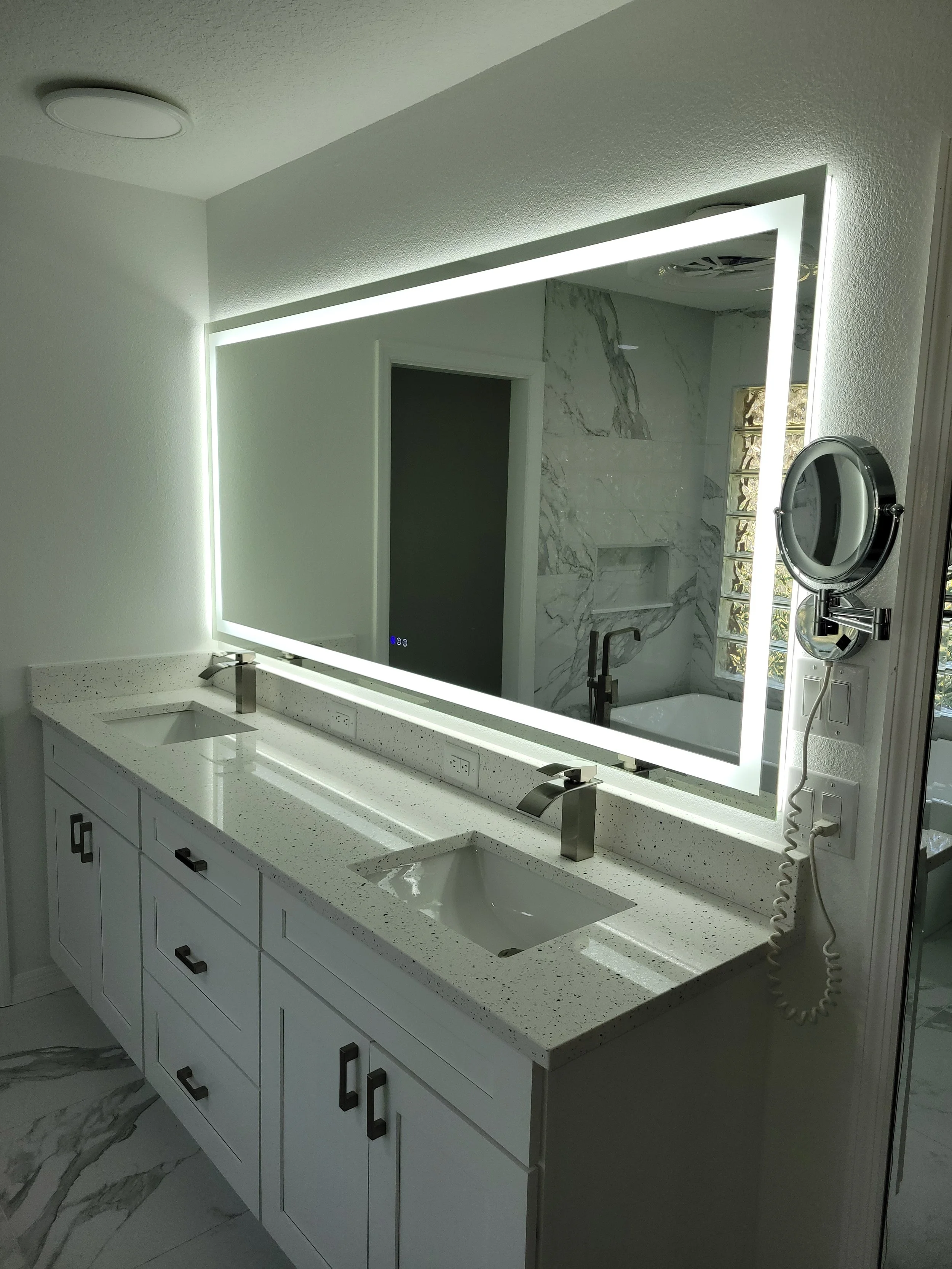 Modern bathroom vanity with white speckled countertop, two integrated sinks, and a large illuminated mirror. The bathroom has marble walls and flooring, and a wall-mounted magnifying mirror.