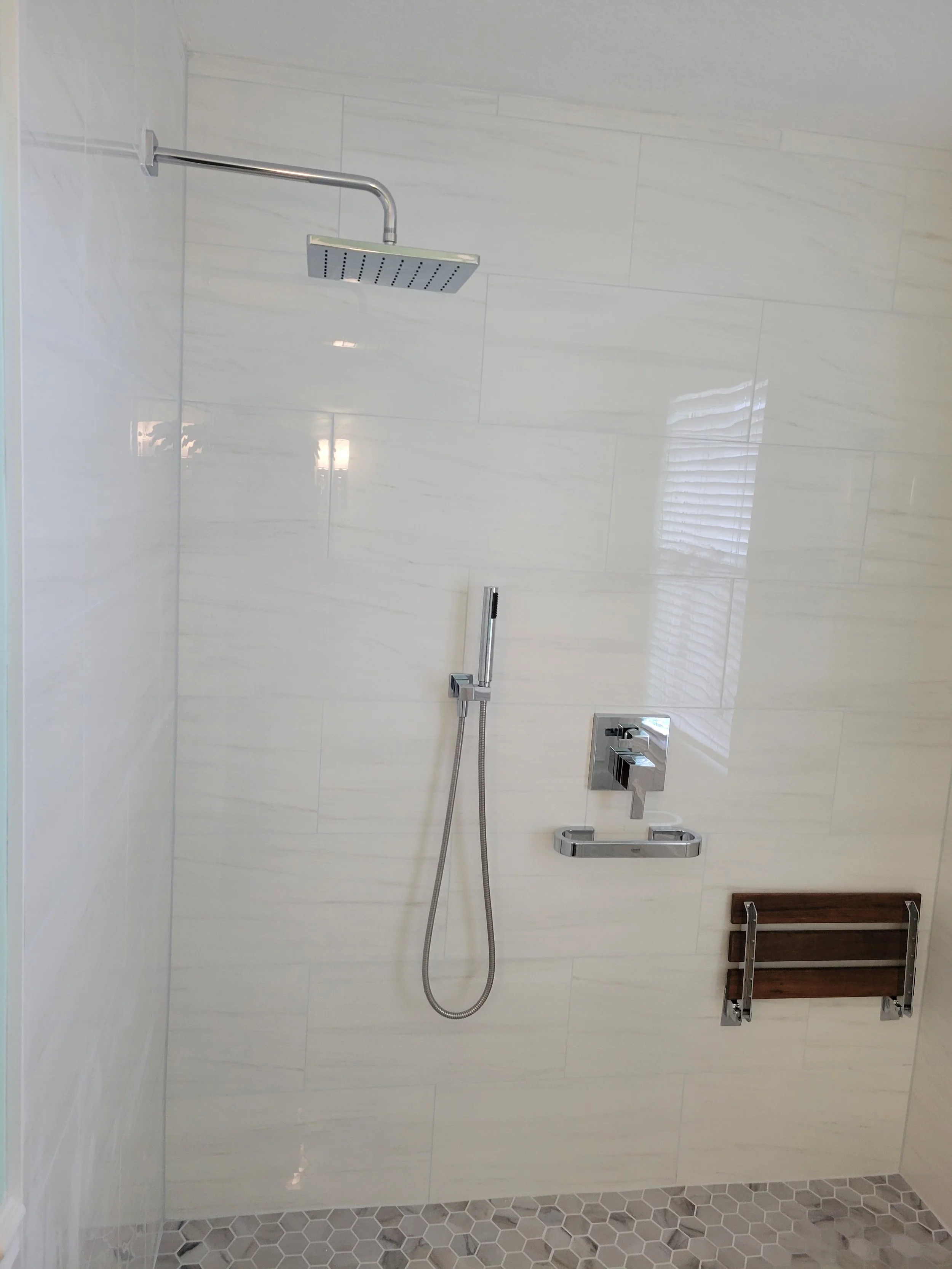 A modern walk-in shower with a rainfall showerhead, a handheld shower, and a wooden foldable shower seat, all mounted on white tiled walls.