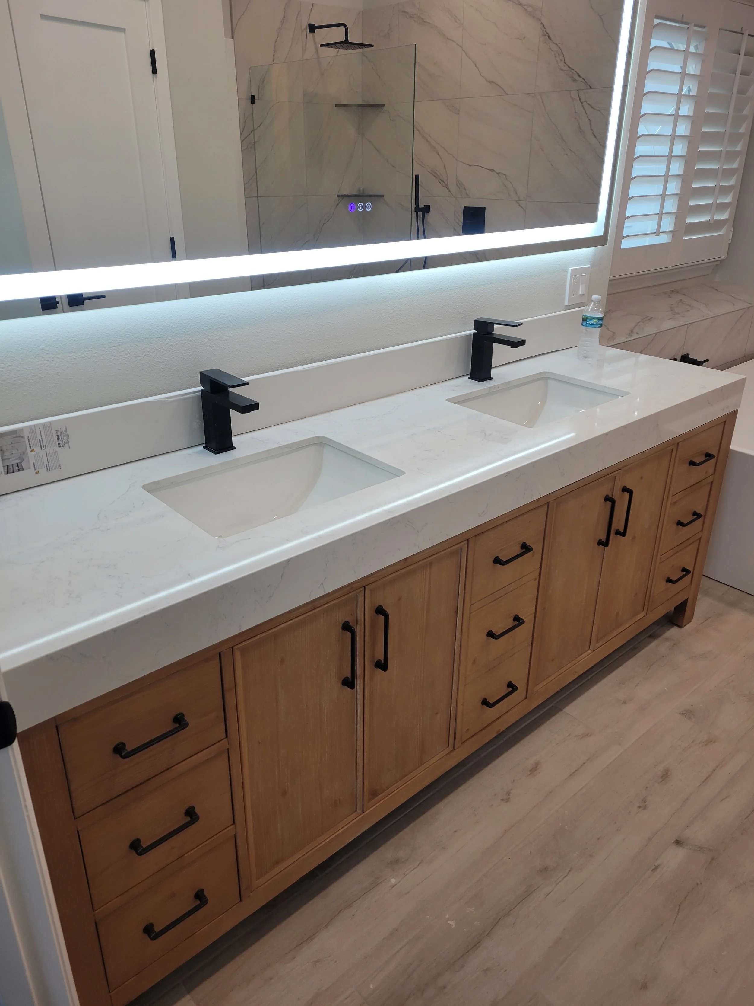 Bathroom vanity with double sinks, black faucets, lots of storage drawers and cabinets, large mirror with backlighting, and a shower with glass door in the background.
