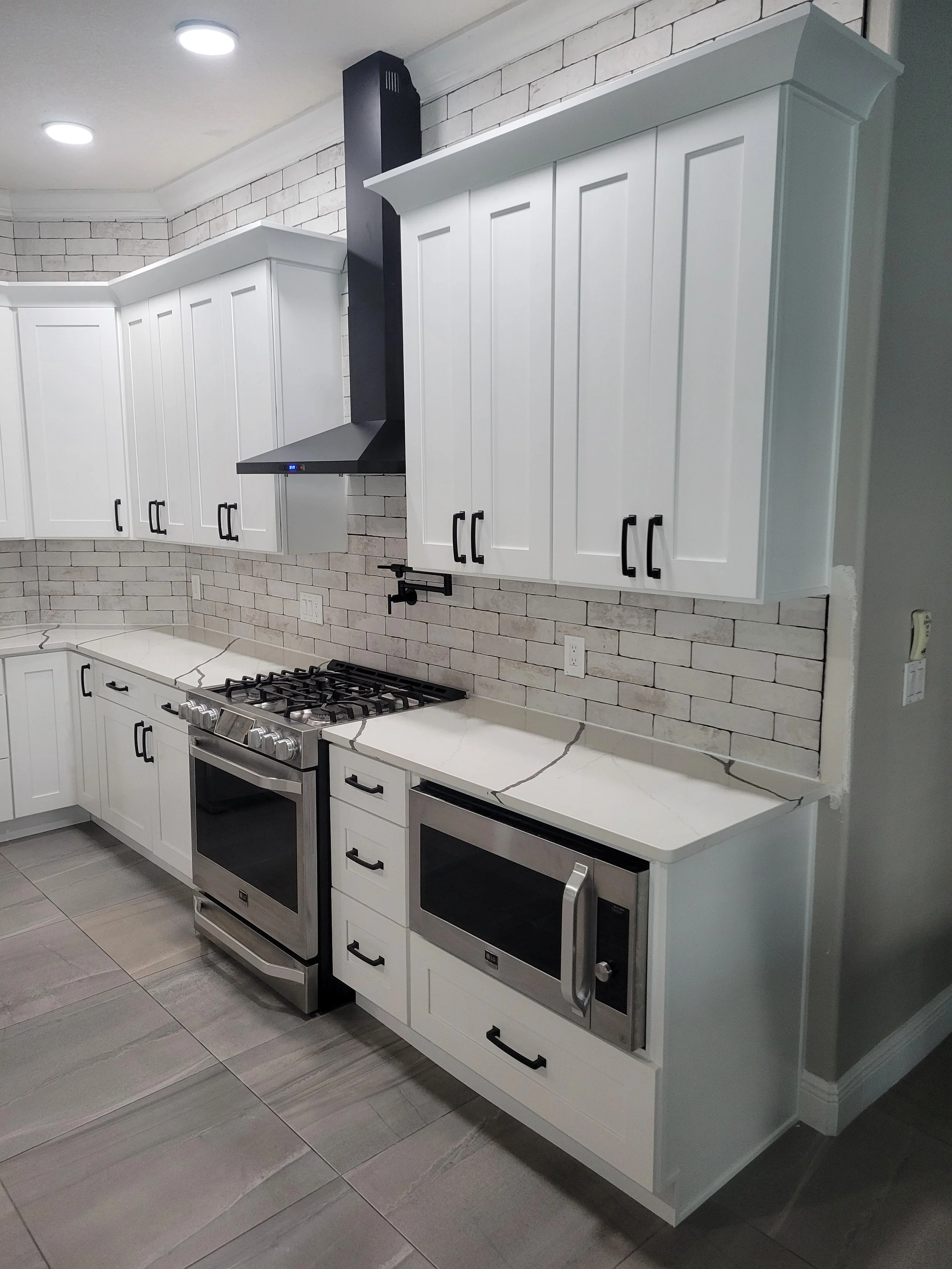 Modern kitchen with white cabinets, black hardware, stainless steel oven and microwave, brick backsplash, and gray tiled floor.