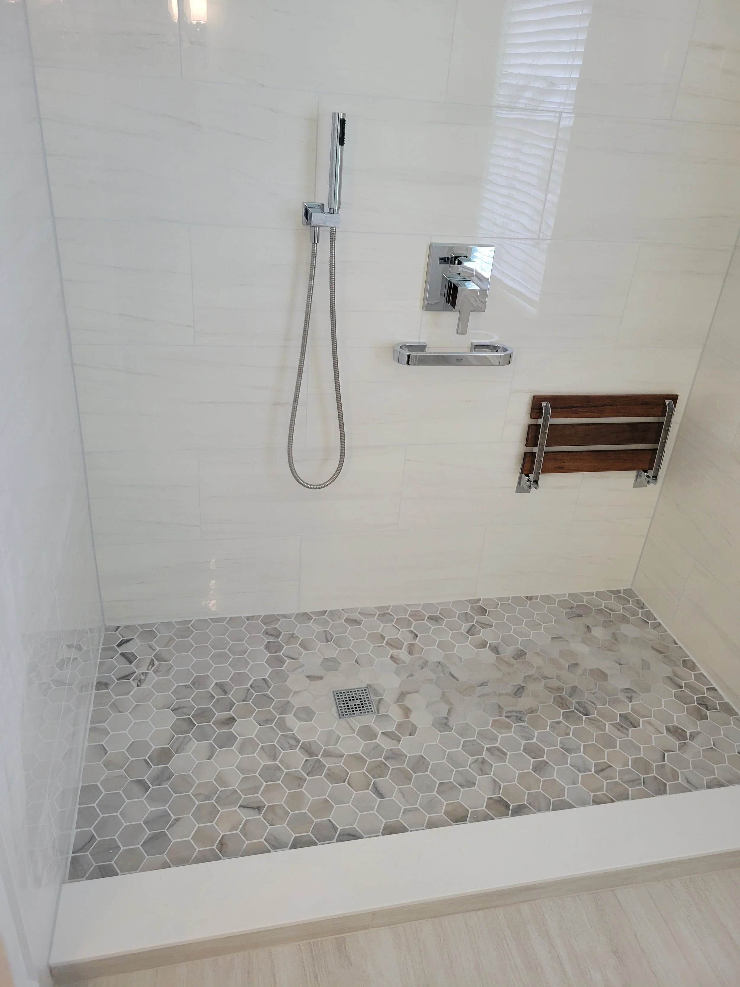 Modern walk-in shower with hexagonal tile floor, a hand-held showerhead on a vertical bar, a chrome faucet with a shelf, and a wall-mounted wooden bench.