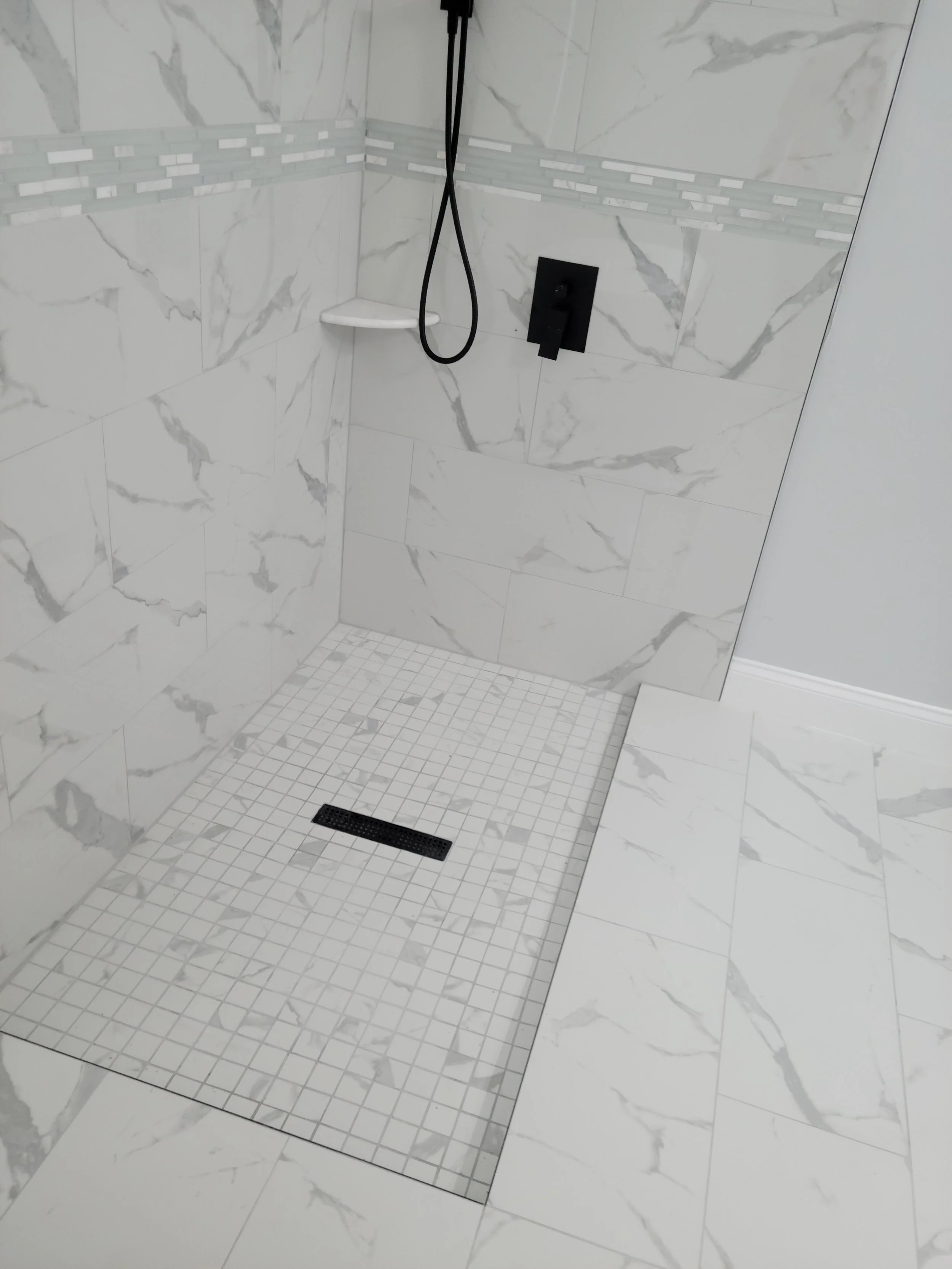 Empty walk-in shower with white marble tiles, a black showerhead, and a narrow shelf in the corner.