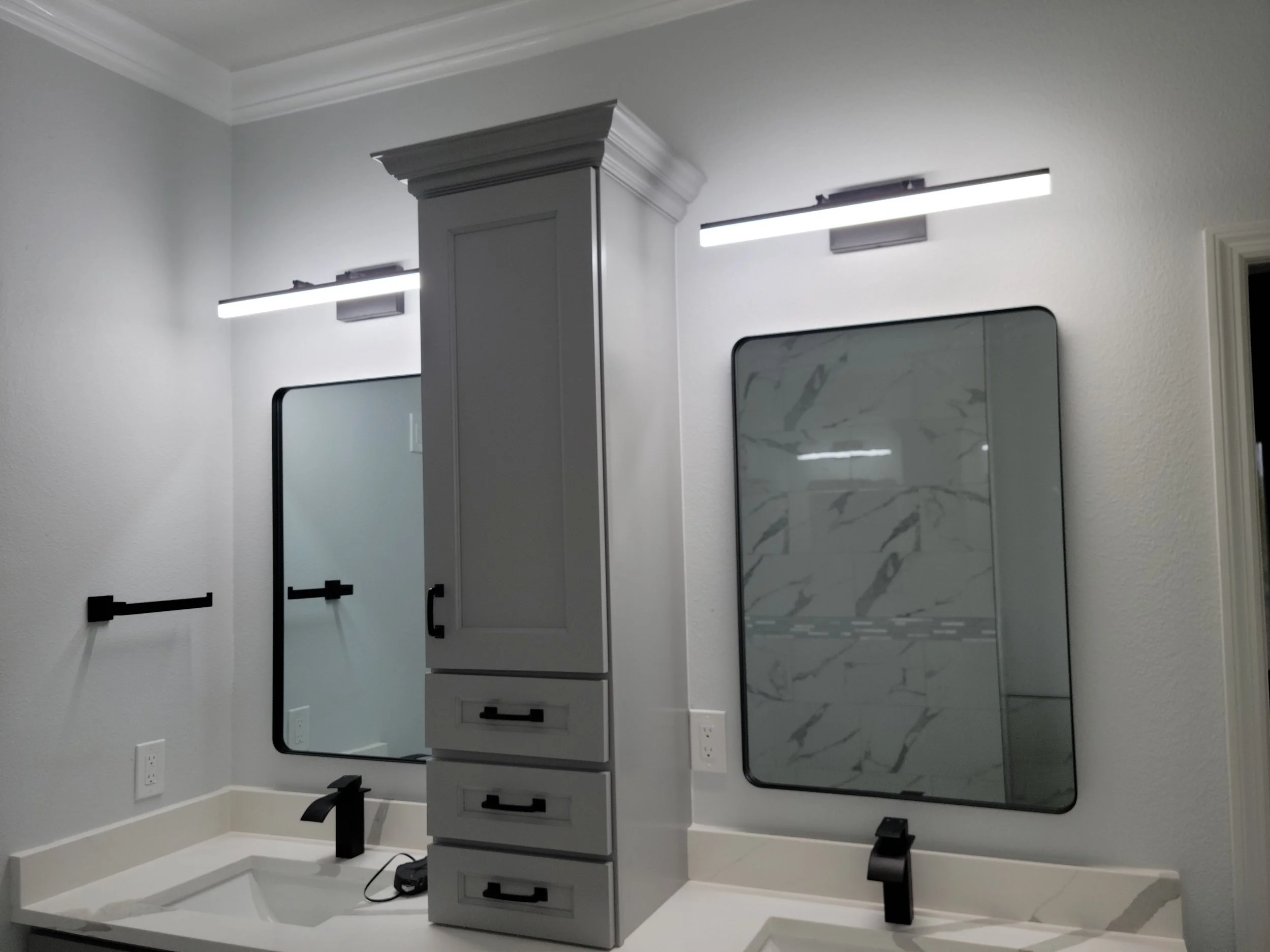 Bathroom vanity with two mirrors, gray cabinets, and two sinks with black faucets, illuminated by modern light fixtures.