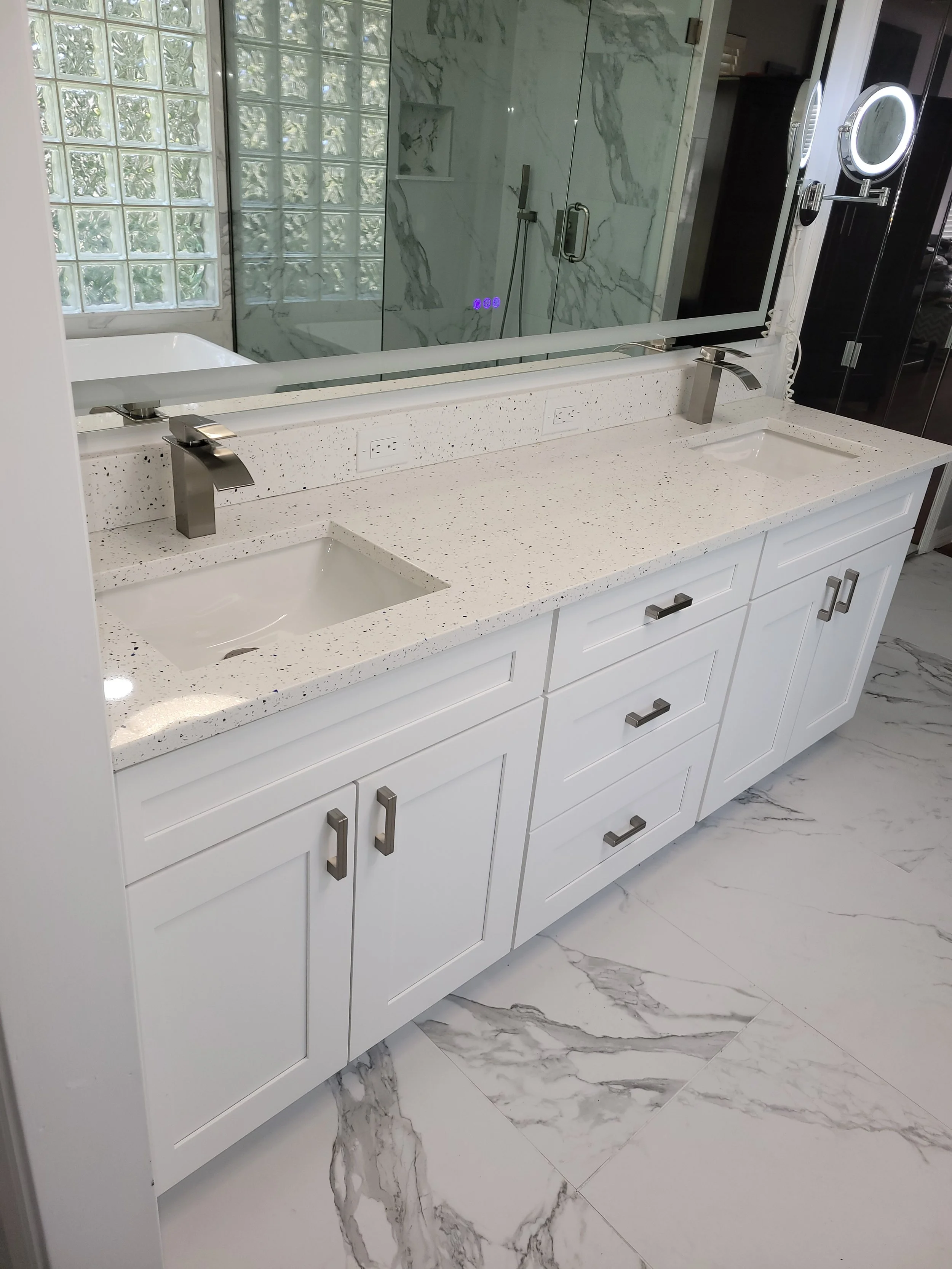 White bathroom vanity with two sinks, a large mirror, and marble flooring, adjacent to a glass-enclosed shower with marble walls.
