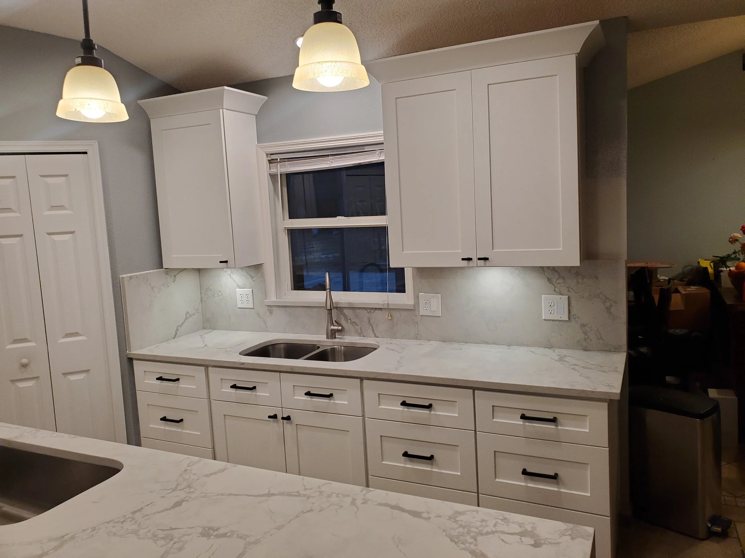 Modern kitchen with white cabinets, marble countertops, dual sink, window above the sink, and overhead pendant lighting.