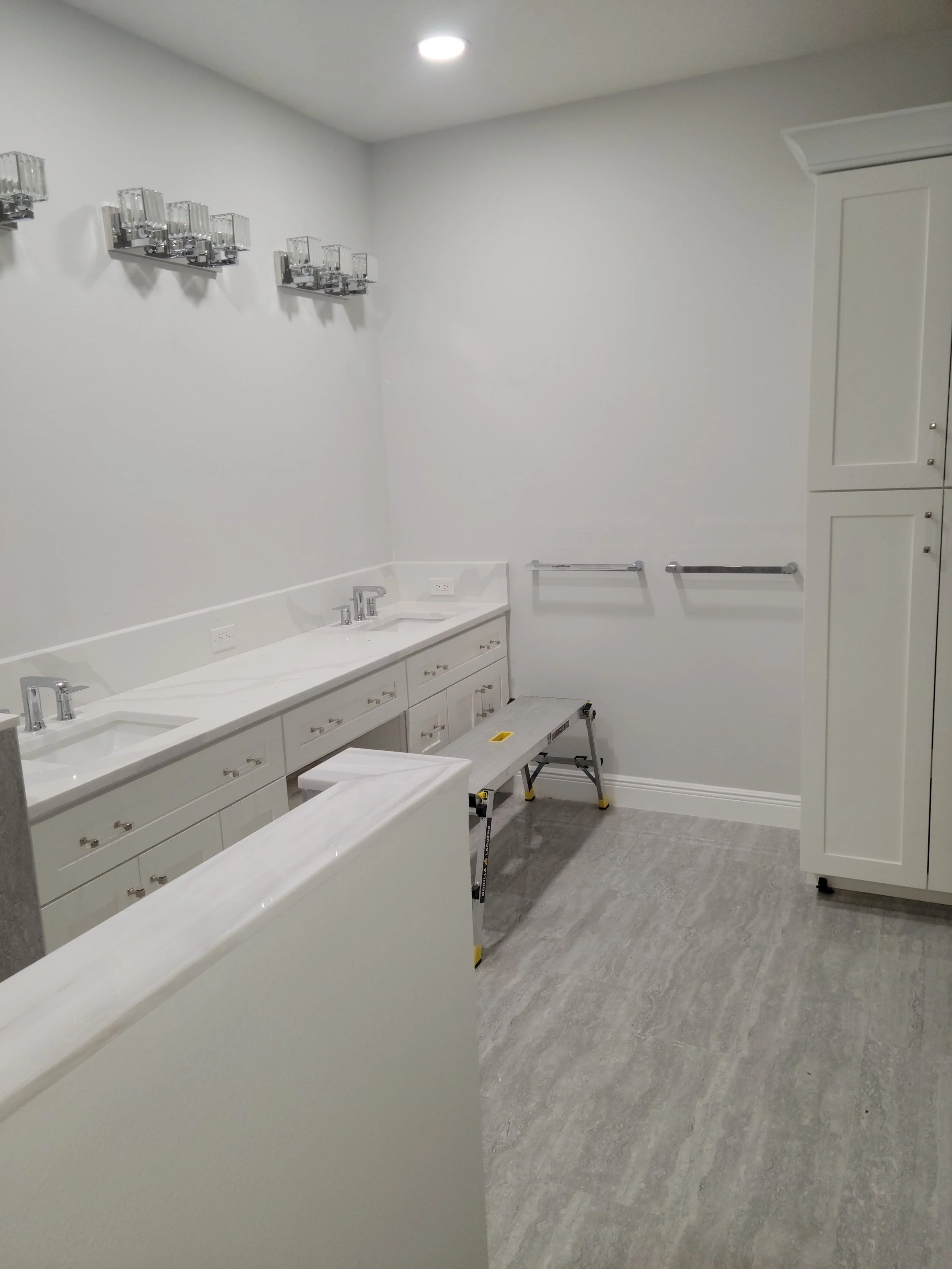 A clean, modern bathroom with white cabinetry, a dual sink vanity, gray floor tiles, wall-mounted lights, and a small step ladder.