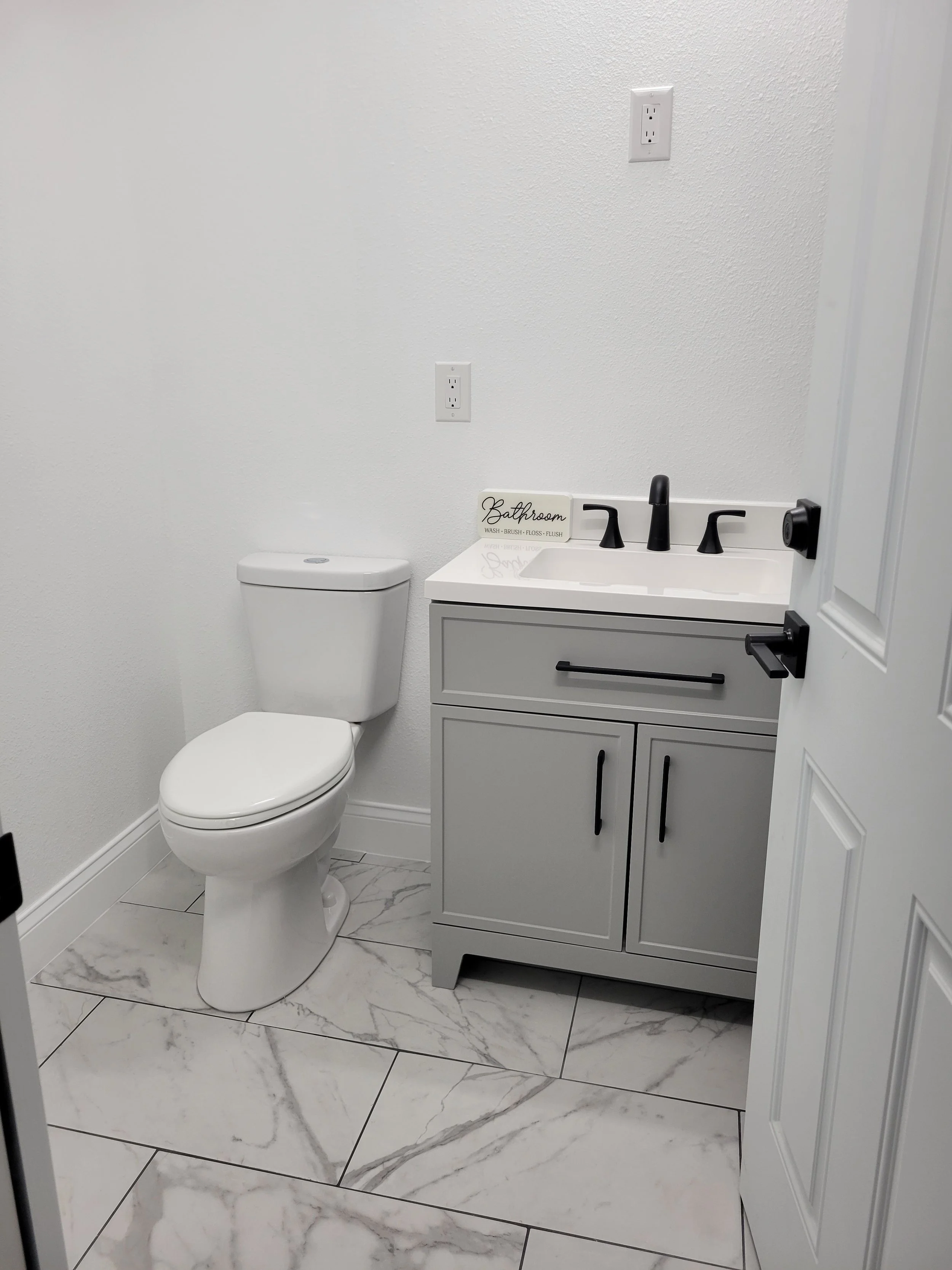 A small bathroom with a white toilet, a gray vanity with black handles, a small white sink, and a black faucet on a white countertop.