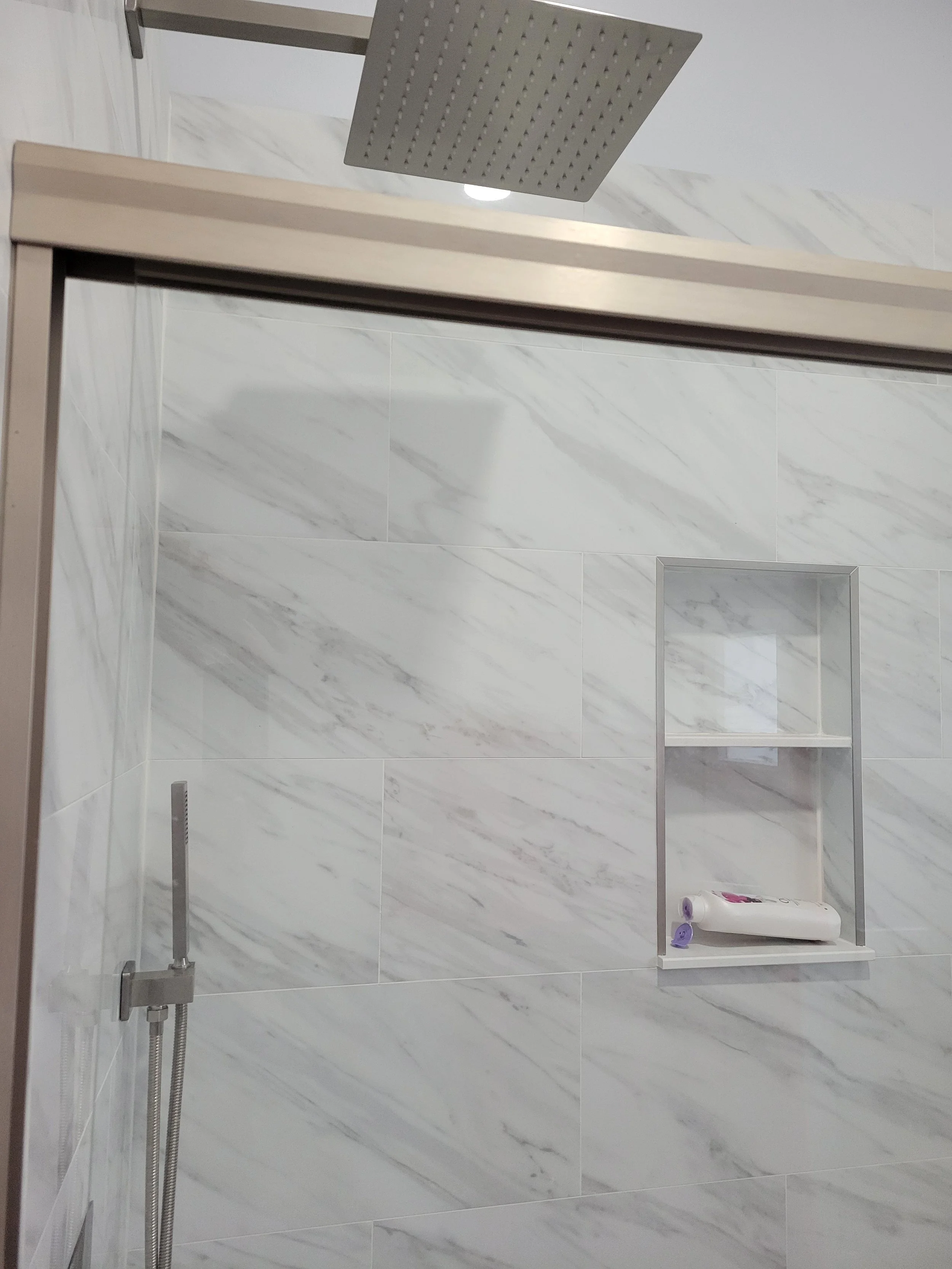 Interior view of a modern shower with white marble tiles, a square rainfall shower head, and built-in wall niche with toiletries.