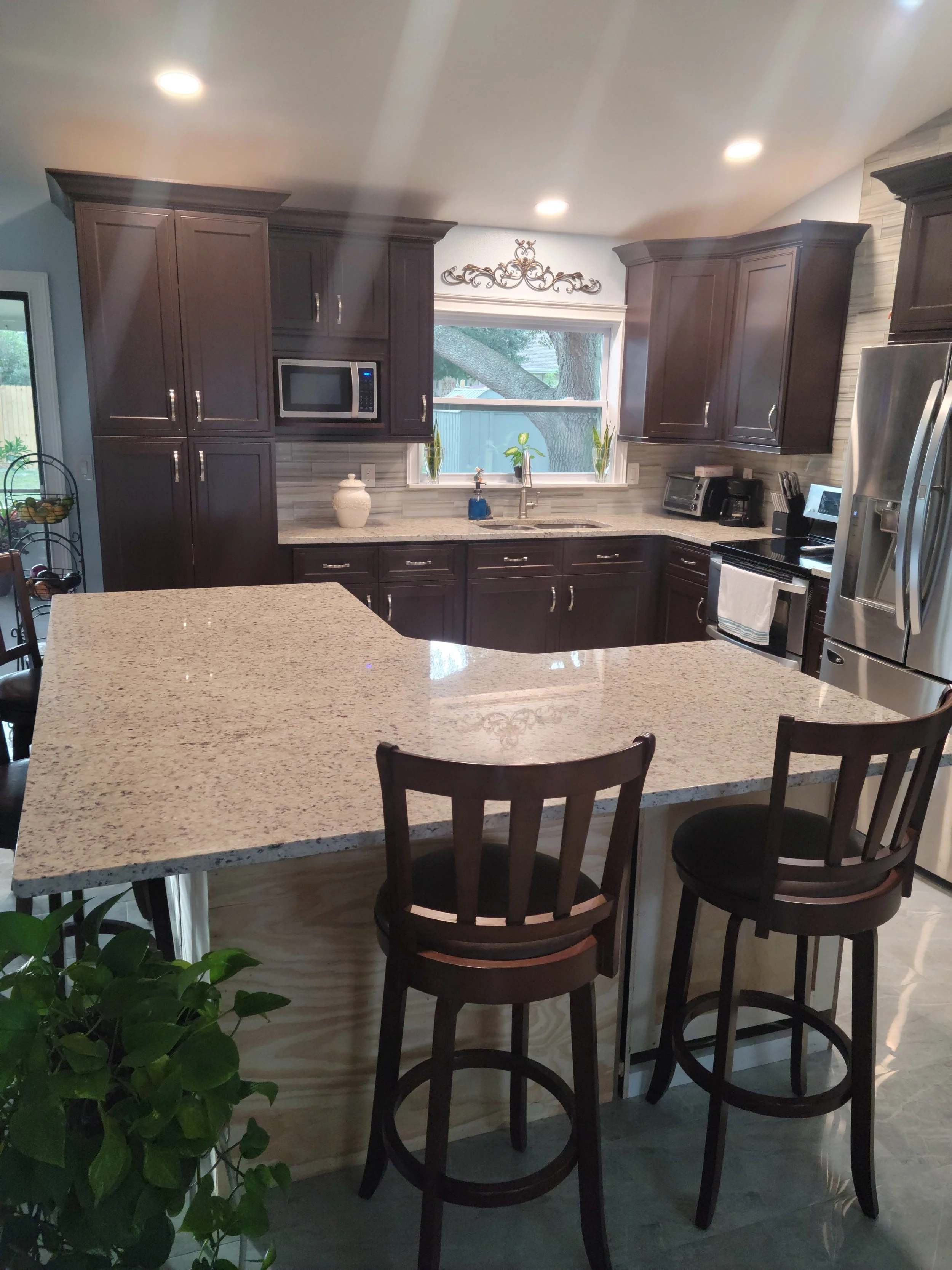 Modern kitchen with dark wood cabinets, granite island, stainless steel refrigerator, microwave, and stove. Window above sink with plants and tree outside.