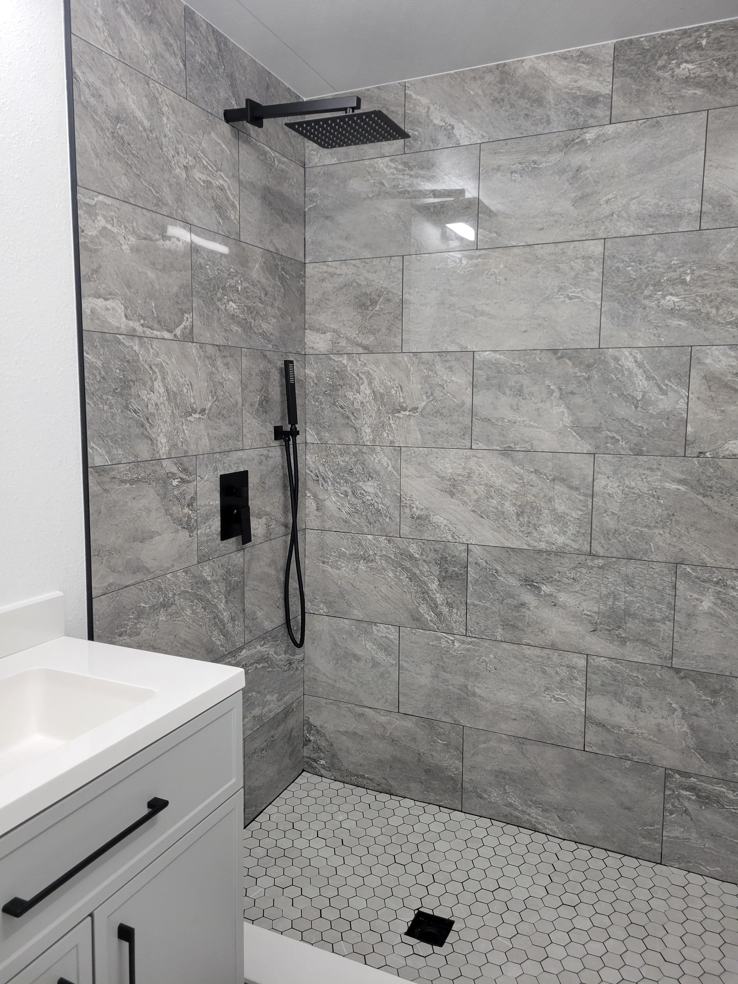Modern bathroom shower area with gray marble tiles, black rainfall showerhead, handheld shower, and floor drain, adjacent to a white vanity with black handles.