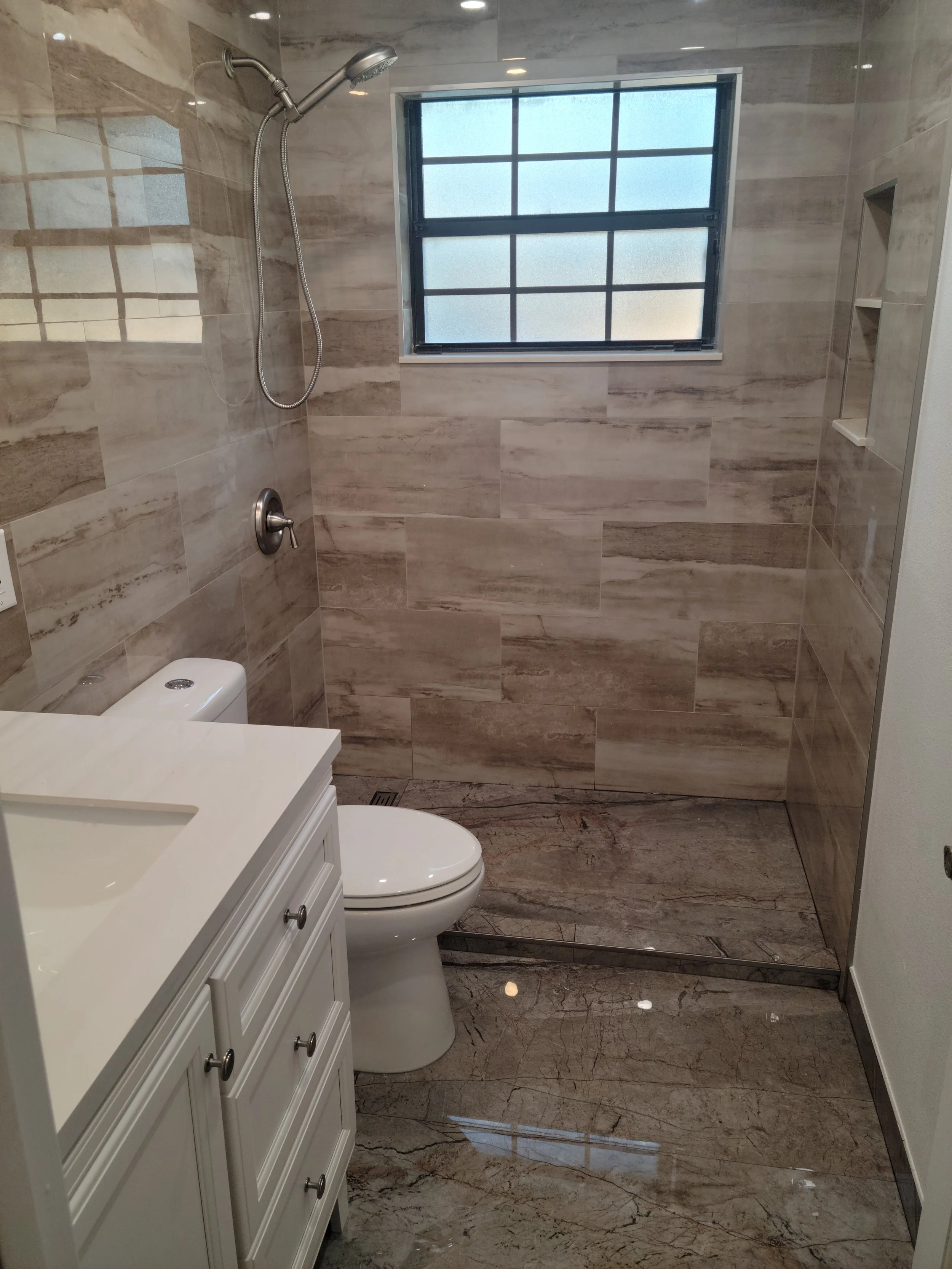 Bathroom with shower area, frosted window, white vanity, and toilet, featuring beige and brown marble tiles.