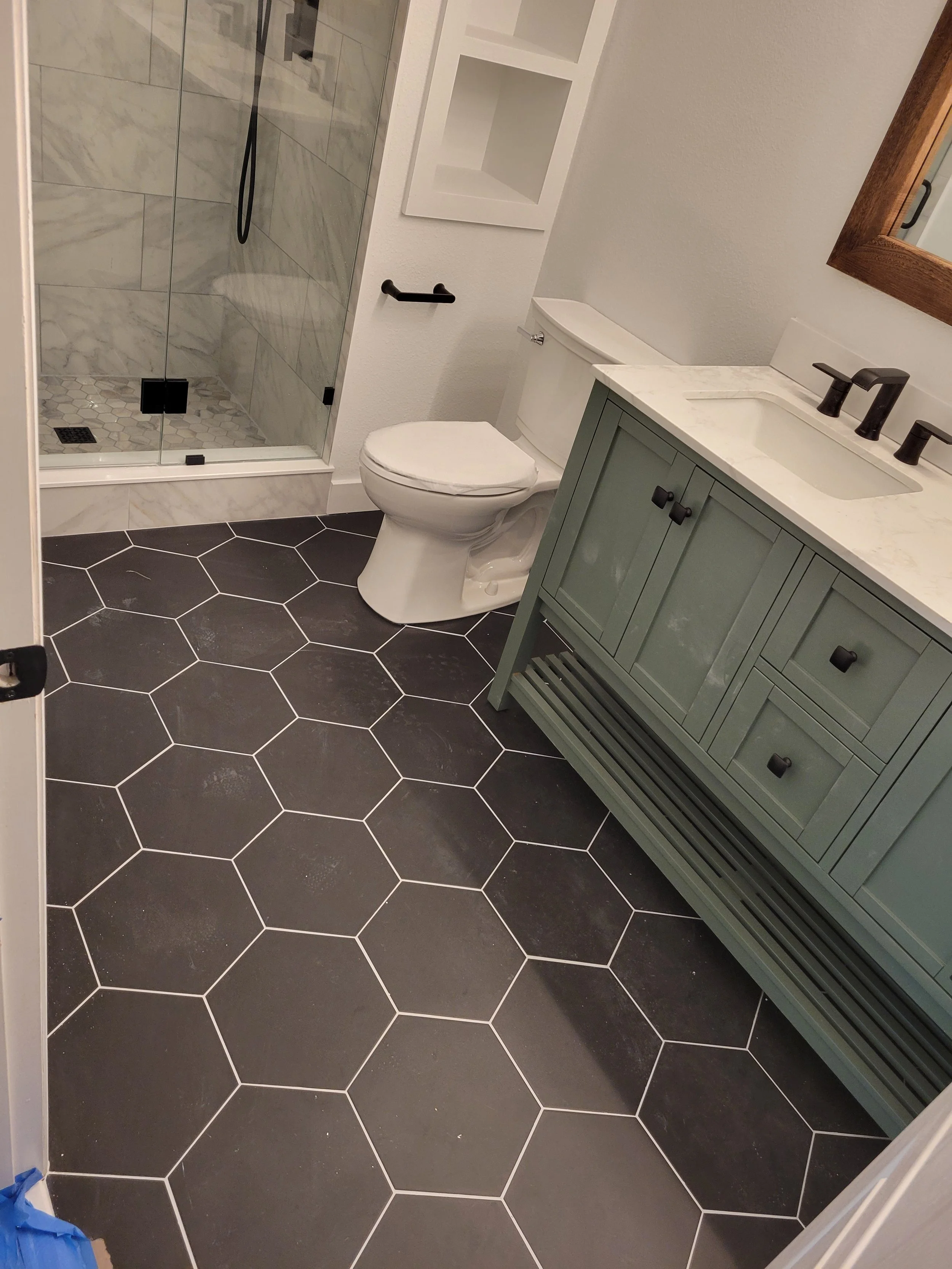 Bathroom with a walk-in shower, a toilet, a light green vanity with a white marble countertop, and black hexagonal floor tiles.