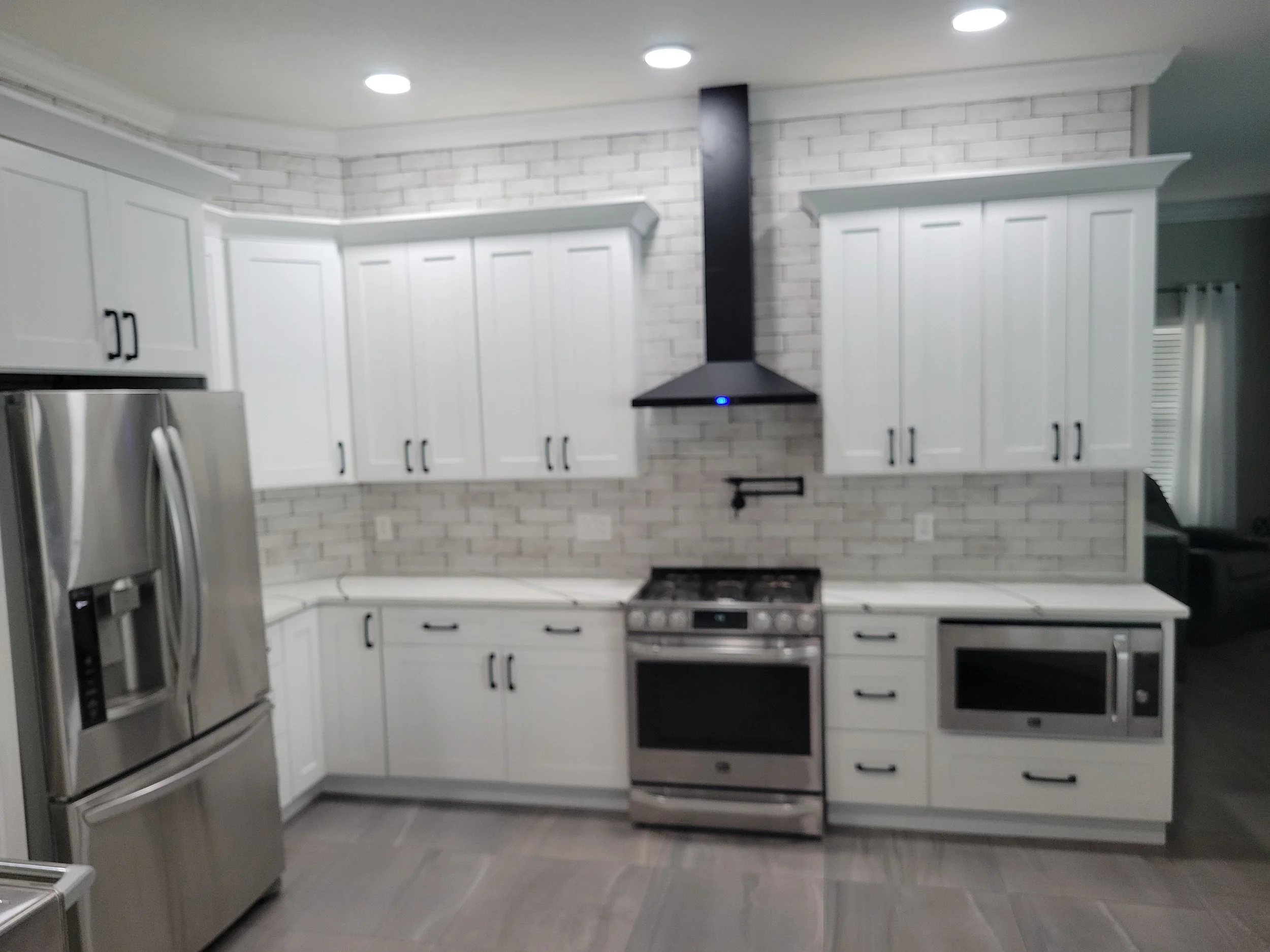 Modern kitchen with white cabinets, stainless steel refrigerator, oven, microwave, black range hood, and gray tile flooring.