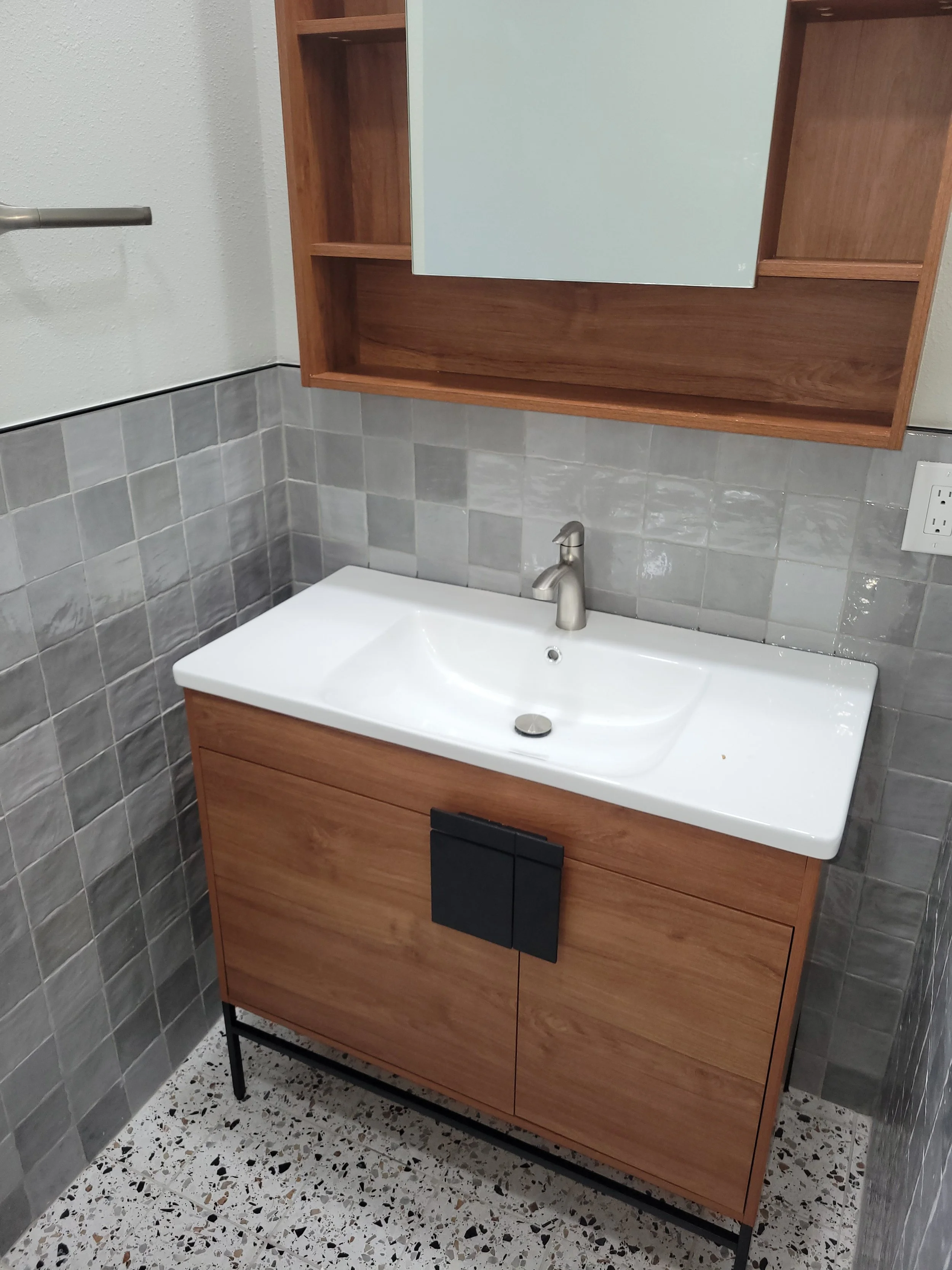 Modern bathroom vanity with a white sink, wooden cabinet, and wall-mounted wooden shelf unit above, with tiled wall and terrazzo floor.